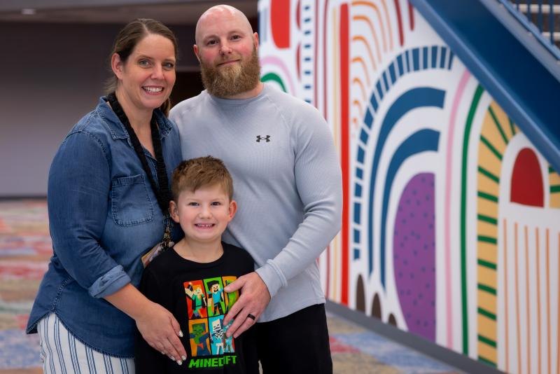 A woman, a man, and a young child pose together in front of a colorful mural.