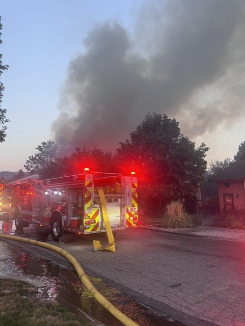 Fire engine with emergency lights on a residential street as thick black smoke rises in the background.