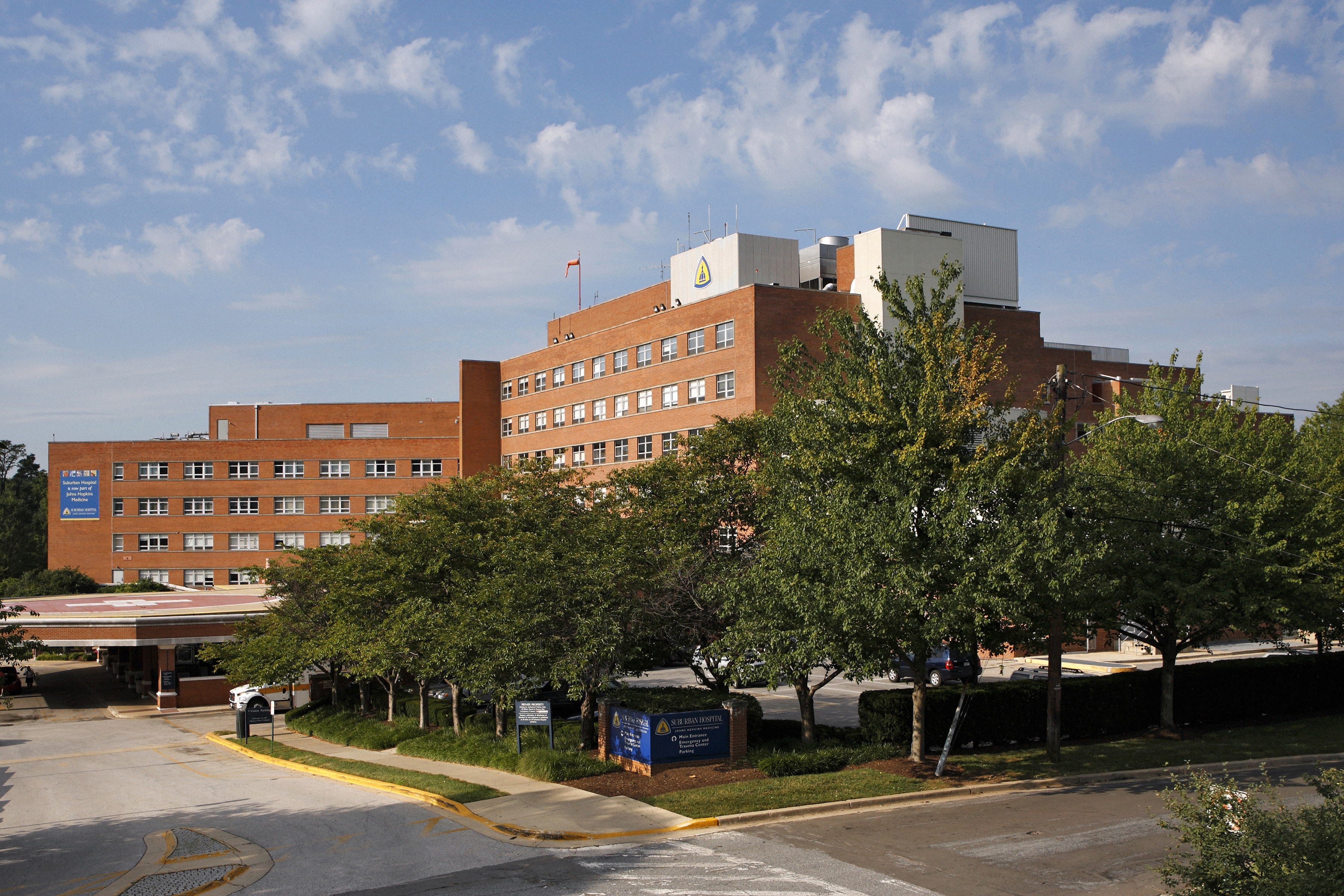 Multi-story red brick hospital building surrounded by mature trees under a blue sky.