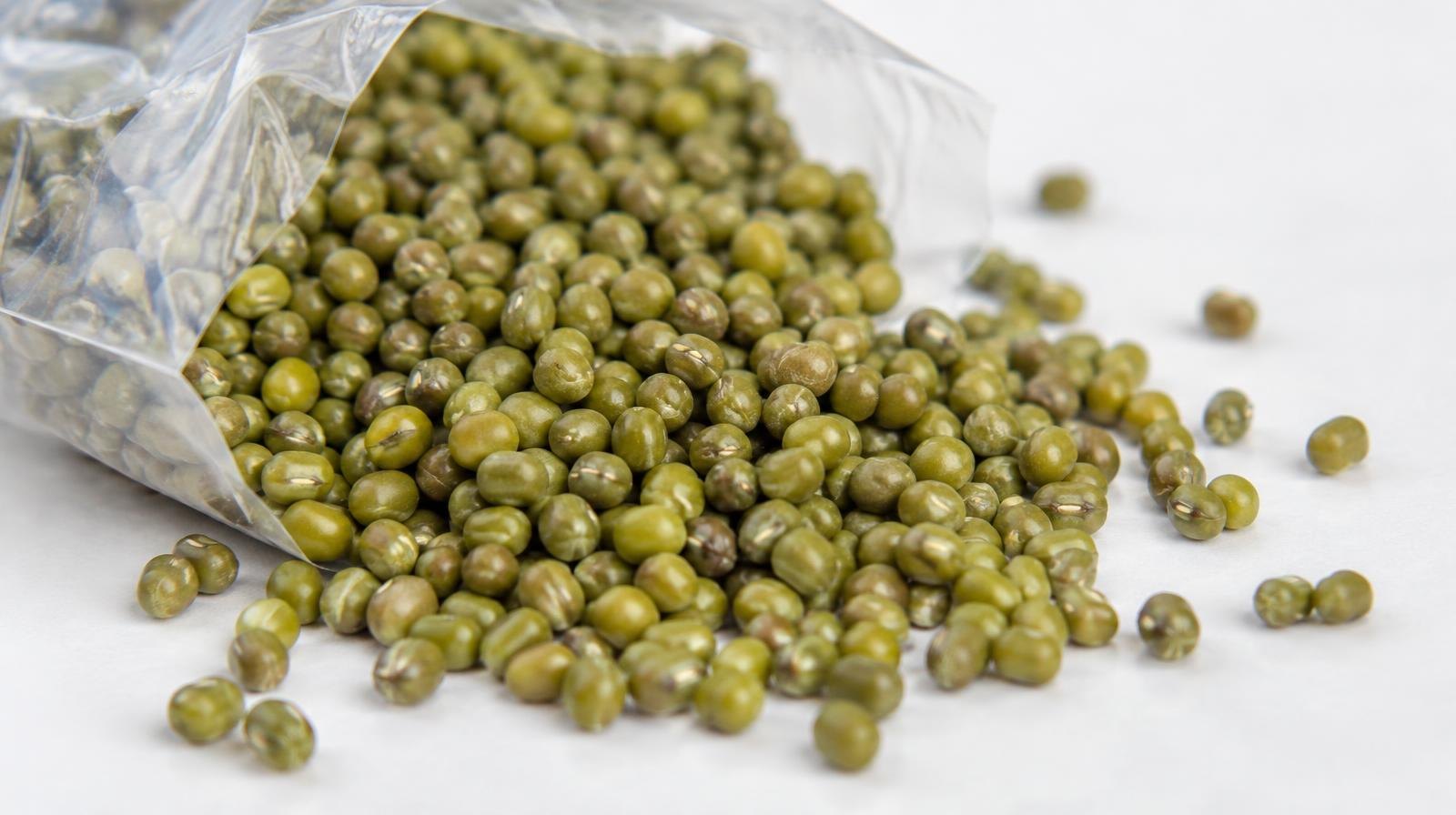 Dried green mung beans spilling from a clear plastic bag onto a white surface.