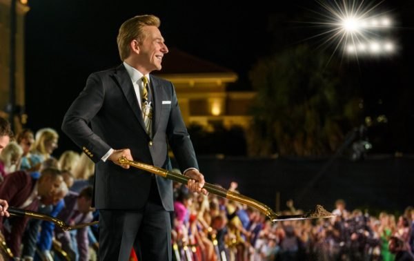 A man in a dark suit holds a ceremonial gold staff on a stage before a large nighttime crowd.
