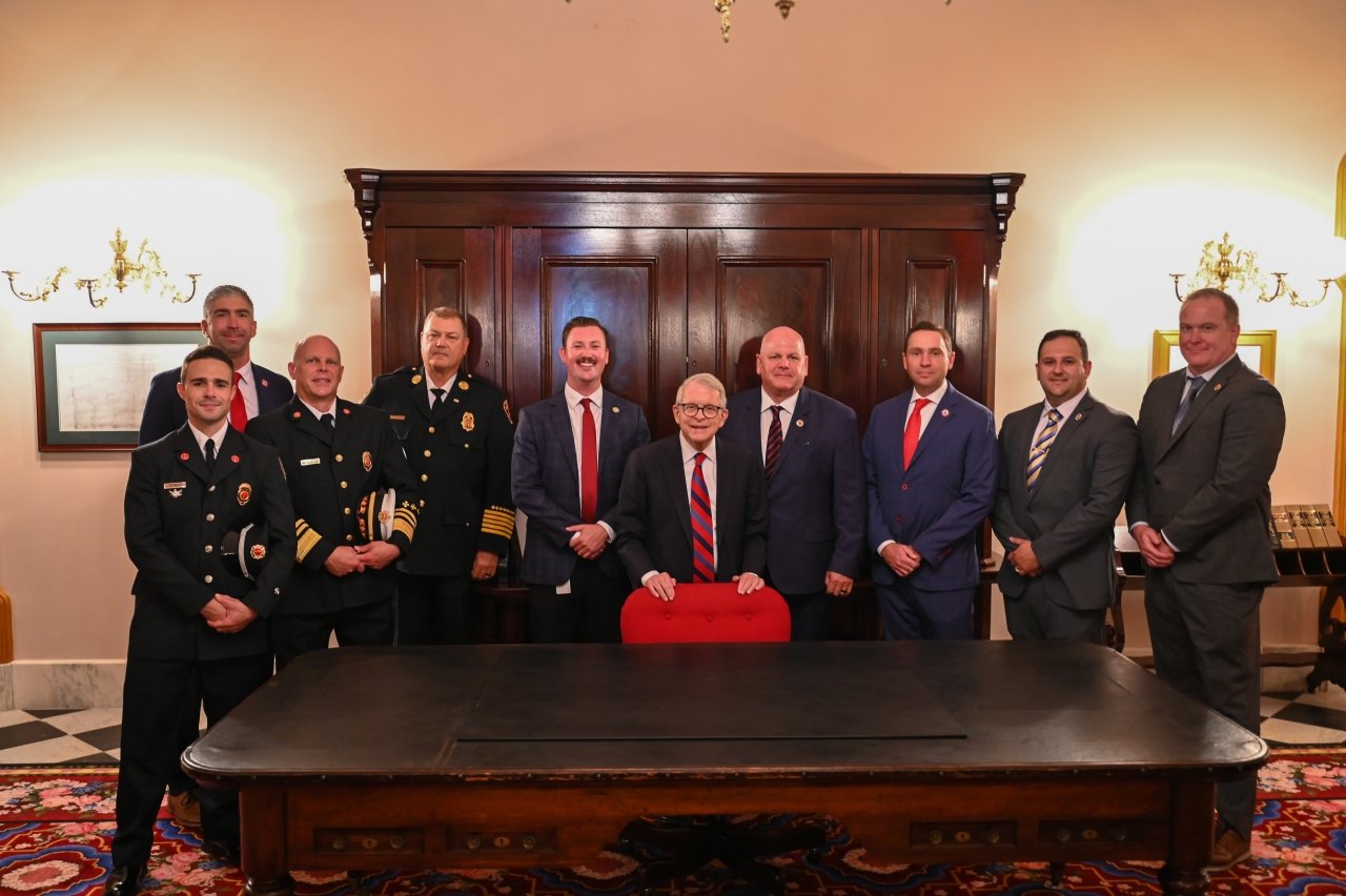 Group of uniformed fire officials and suited officials gathered around a desk during a bill signing.