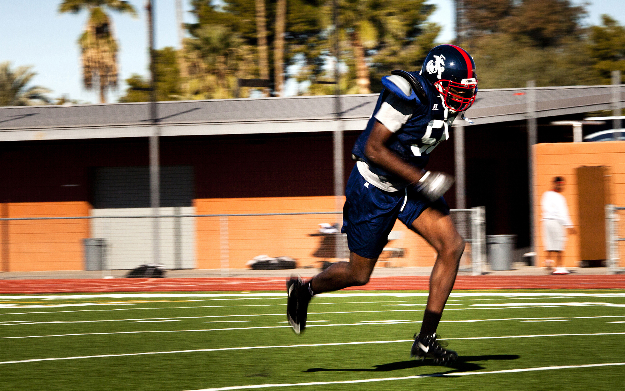 Football player in navy uniform and red helmet sprinting across a grass field.