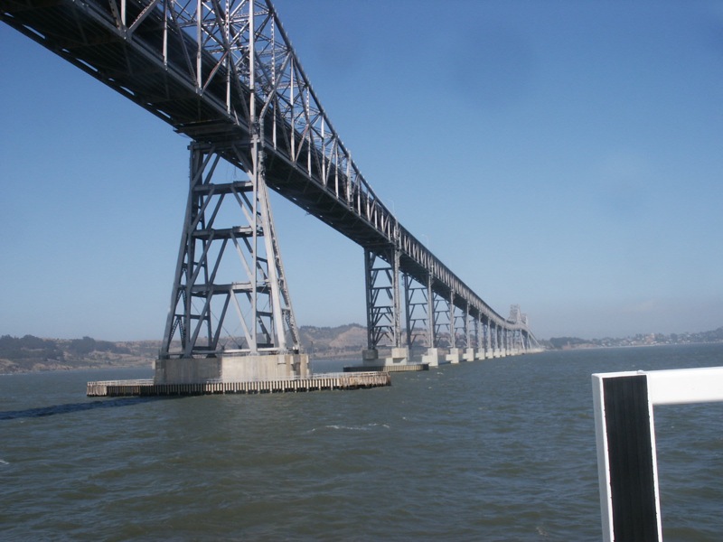 Steel truss bridge spanning a bay viewed from water level under a clear blue sky.