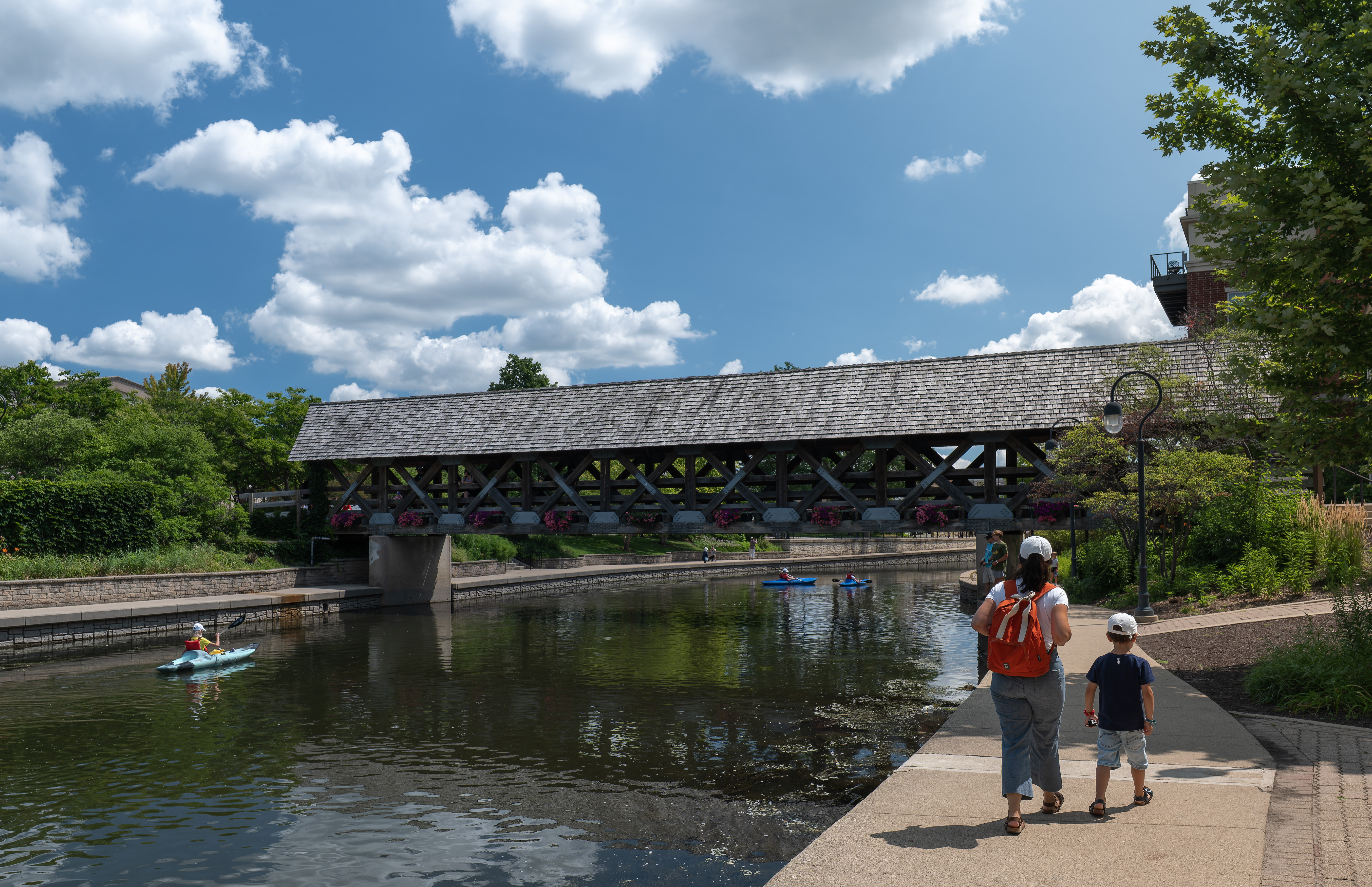 Two people stroll along a paved riverside path near a wooden covered bridge.