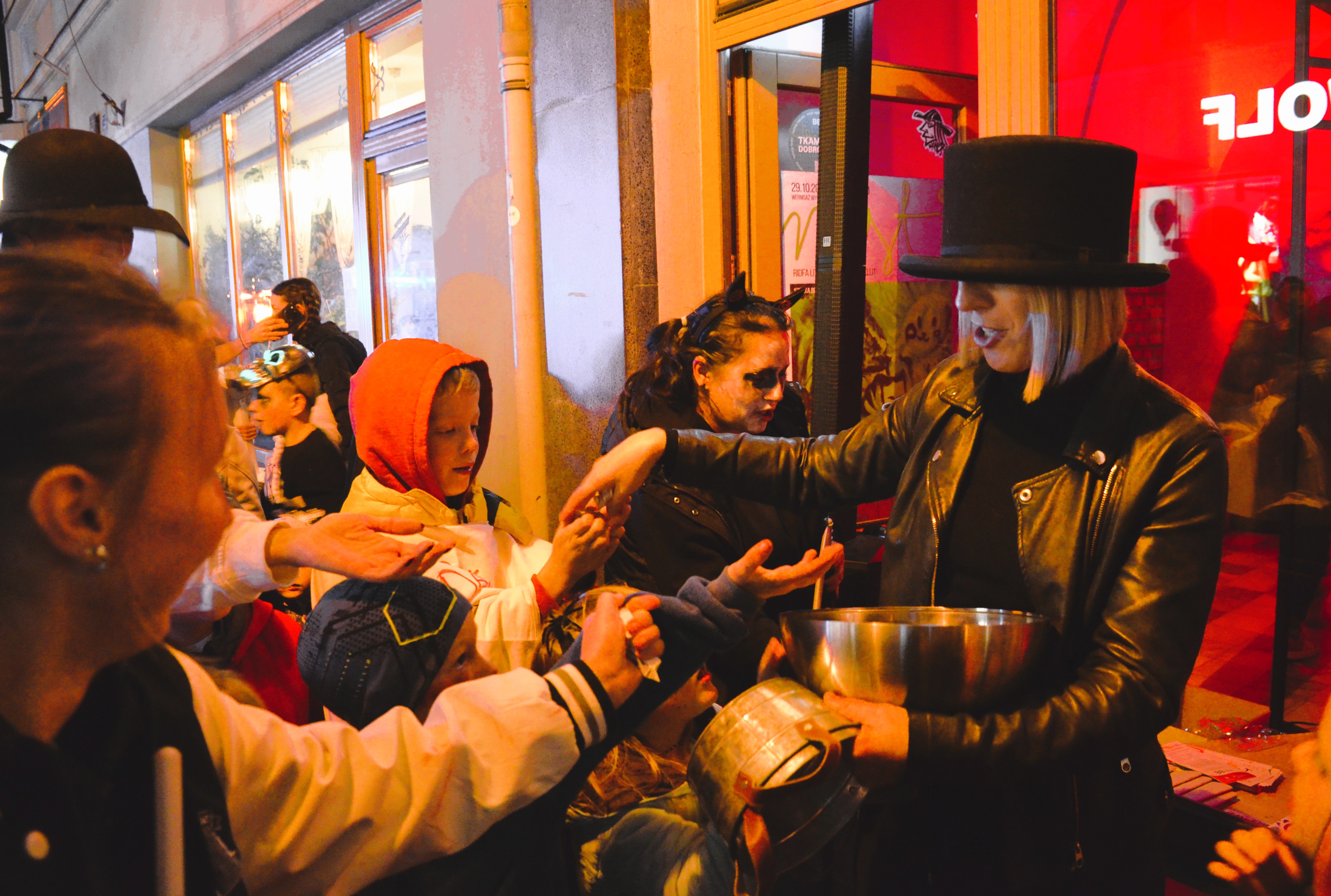 Costumed children reach for treats from an adult holding a bowl on a lit street at night.