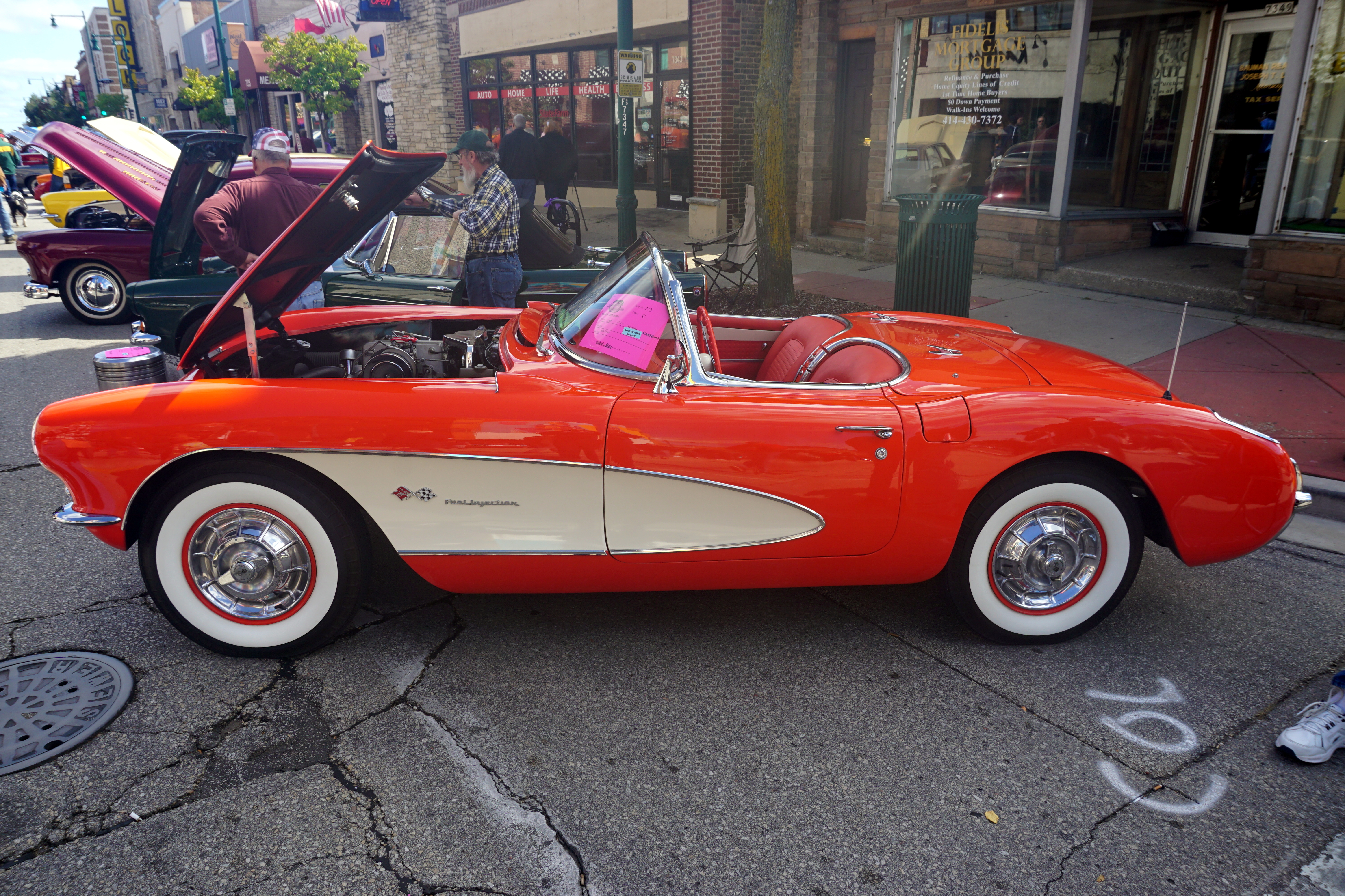 Red and white 1957 Chevrolet Corvette convertible with whitewall tires at an outdoor car show.