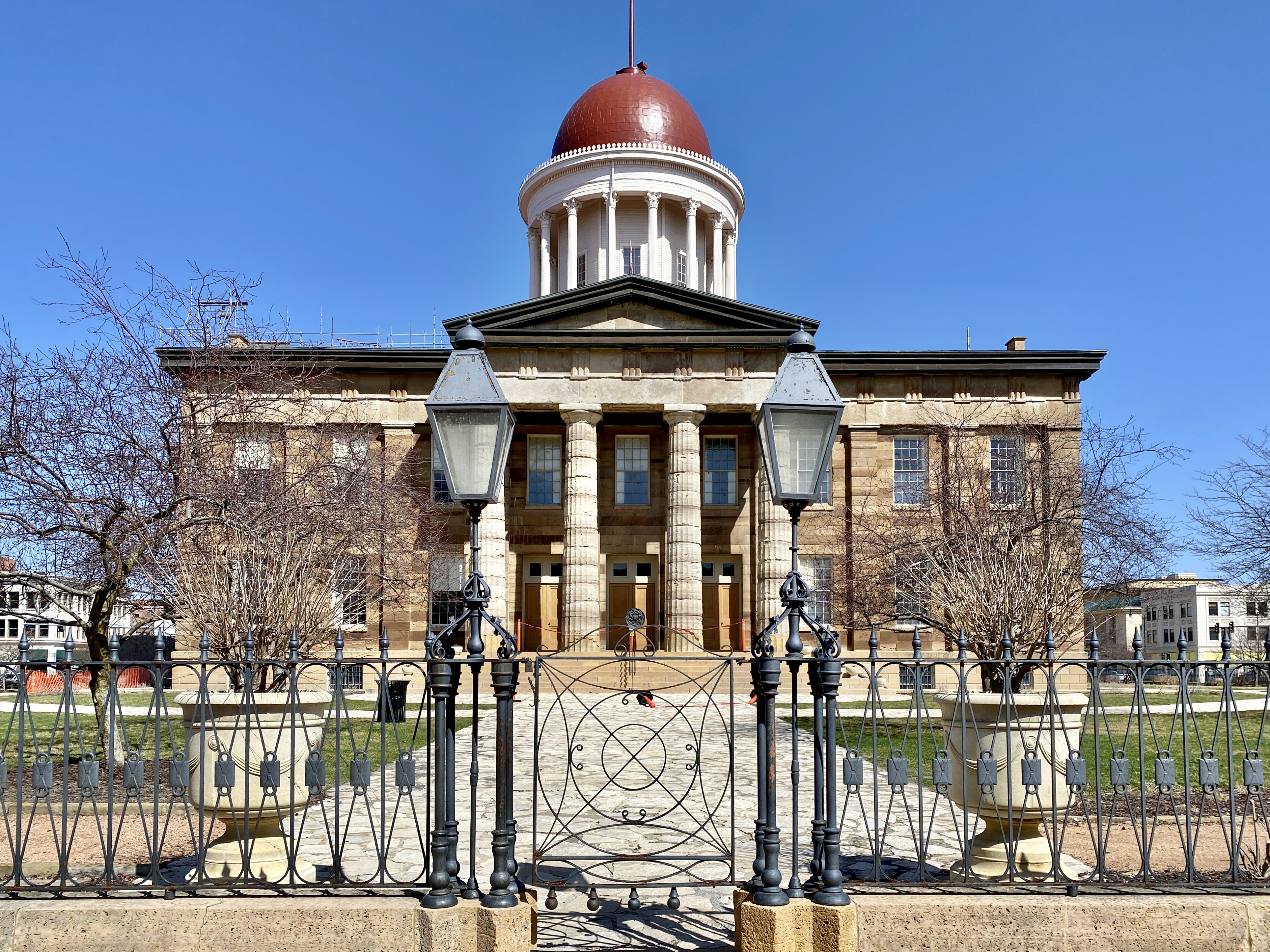 Greek Revival limestone government building with red dome and columned portico under a clear sky.