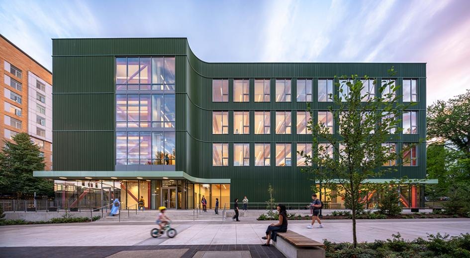 Four-story green metal-clad building with large windows and pedestrians in the foreground plaza at dusk.