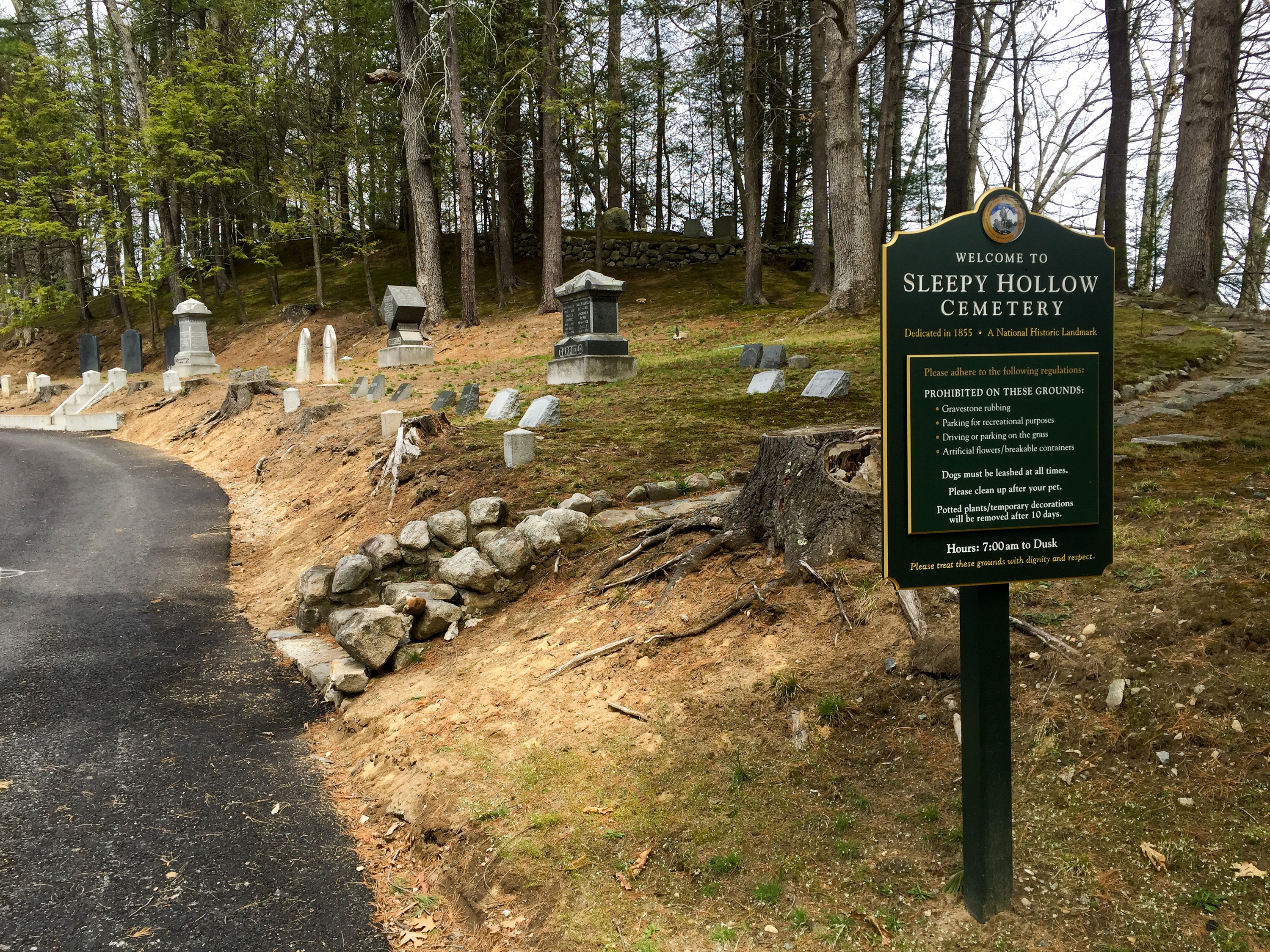 Green welcome sign for Sleepy Hollow Cemetery along a paved path, with gravestones and trees on a hillside behind it.