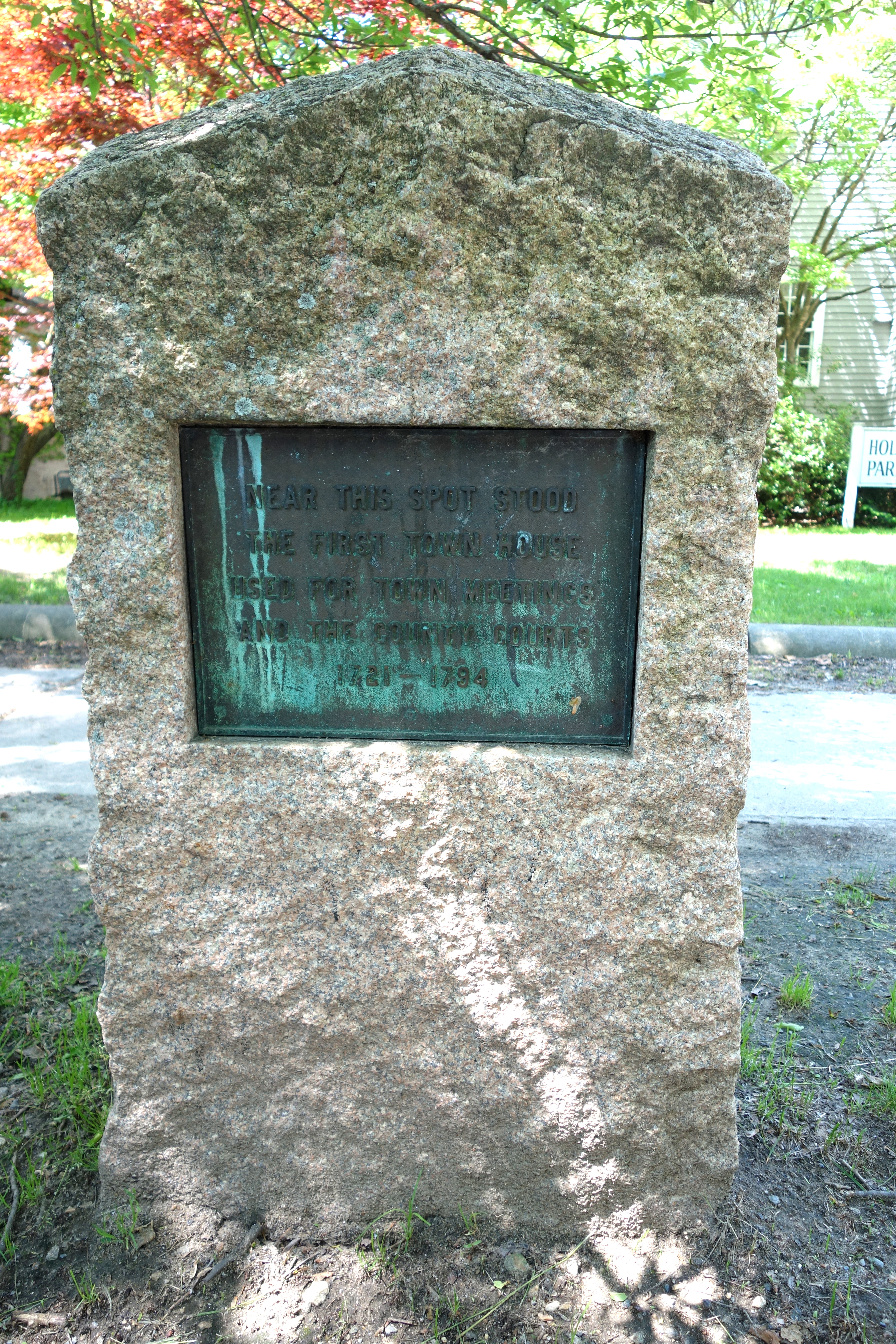 Granite memorial stone with bronze plaque stands outdoors among trees.