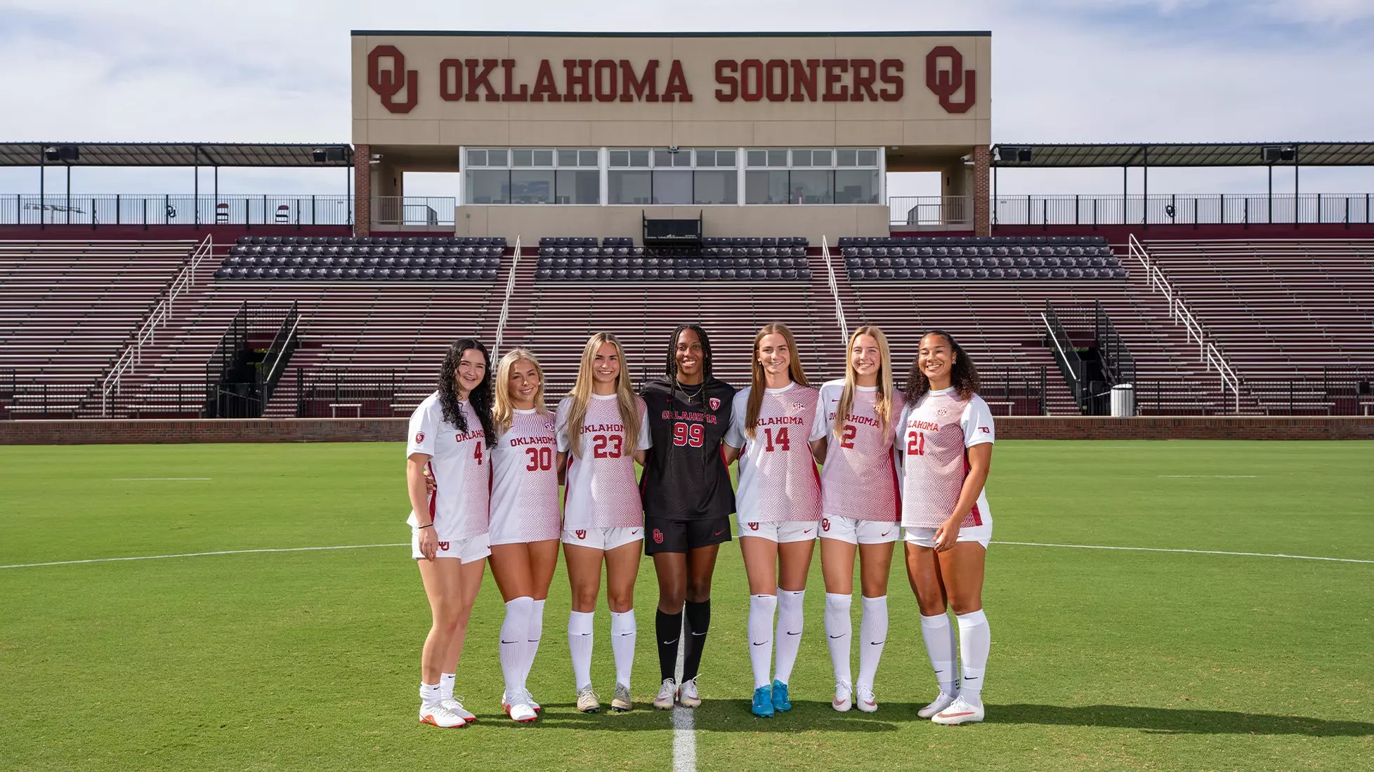 Seven soccer players in Oklahoma Sooners uniforms posed on a field in front of stadium signage.