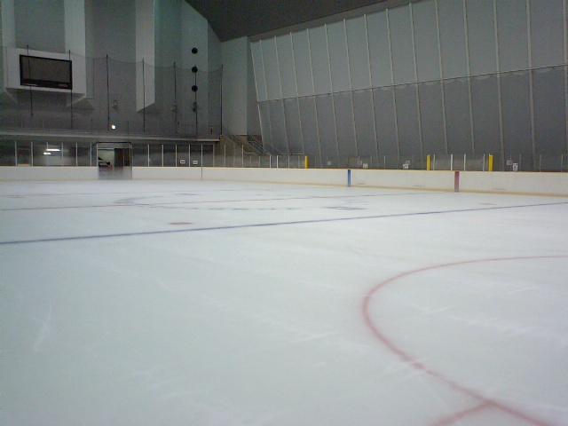 Empty indoor ice rink with white ice surface, red circle markings, and dasher boards.