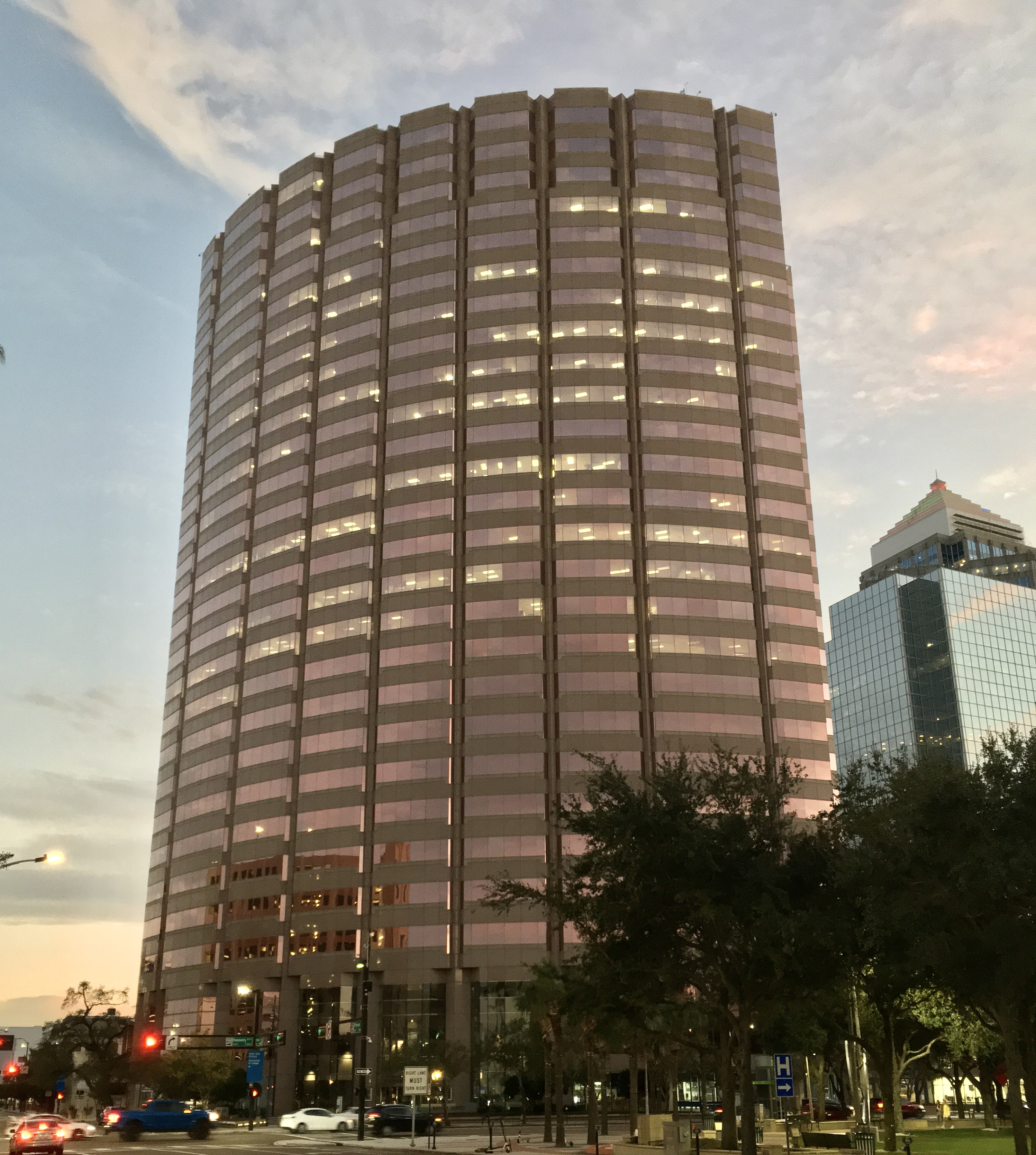 Tall cylindrical government office tower at dusk with trees in the foreground.
