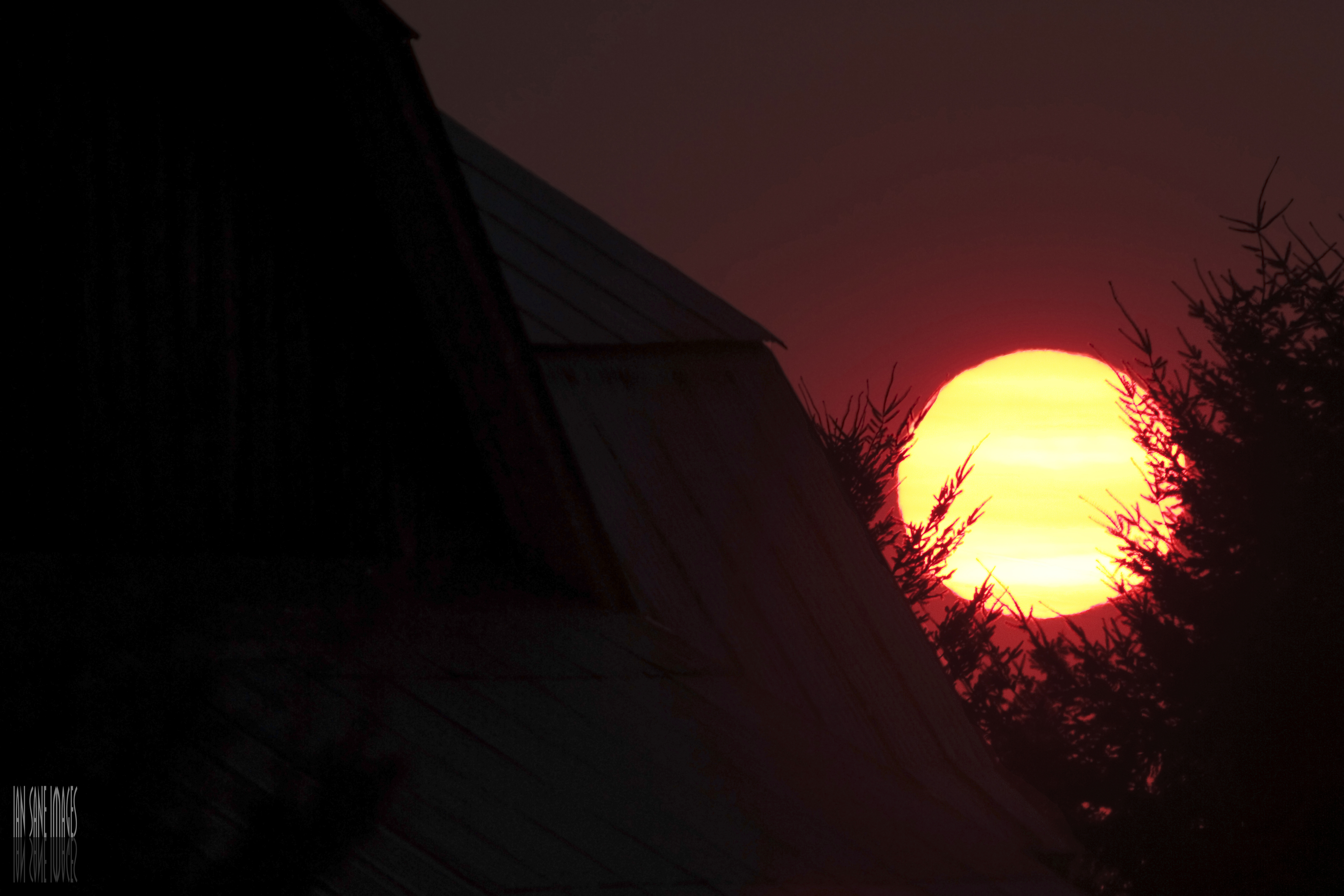 Deep-red sun setting behind silhouetted trees and a barn, sky tinted crimson by wildfire smoke.