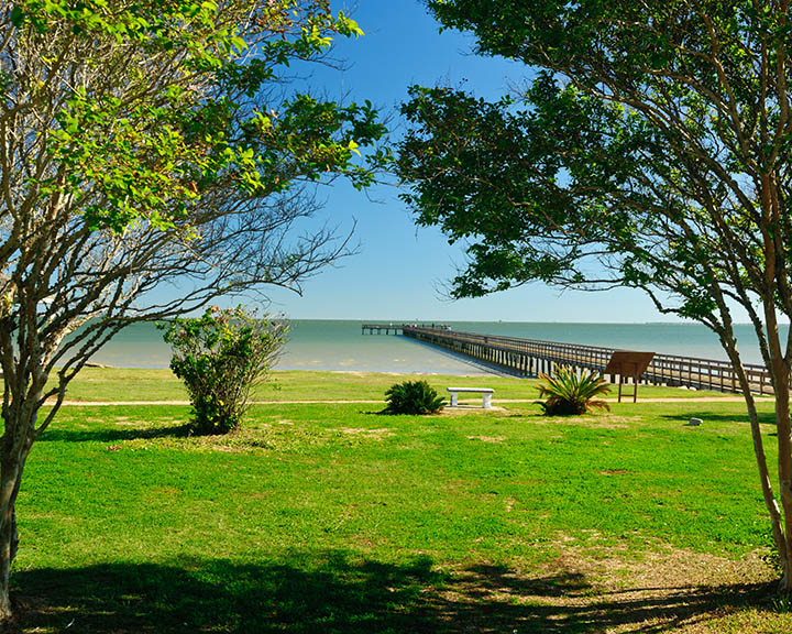 A long fishing pier extends into Galveston Bay from a grassy park with trees.