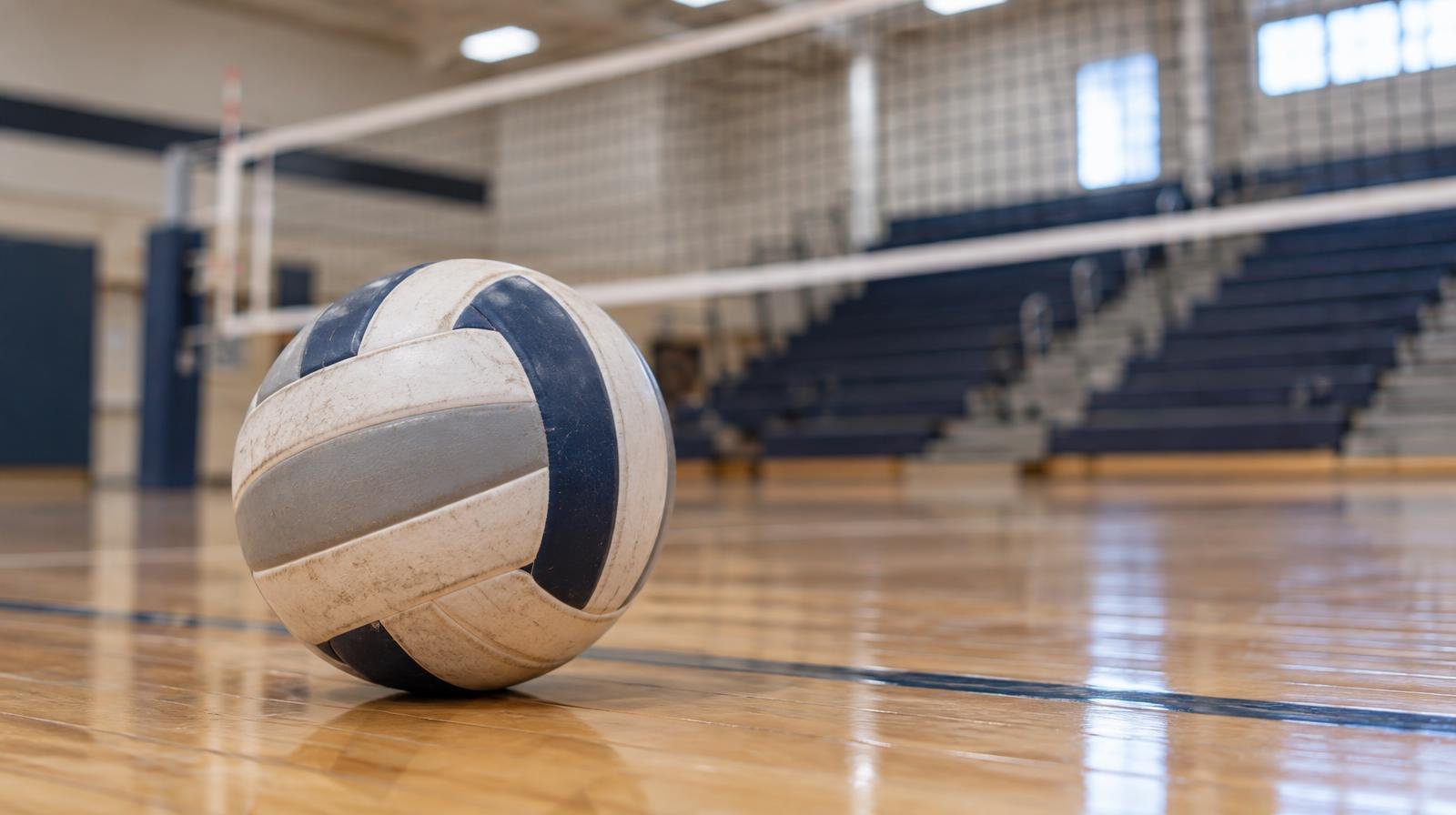 A volleyball resting on a hardwood gymnasium floor near a net with bleachers in the background.