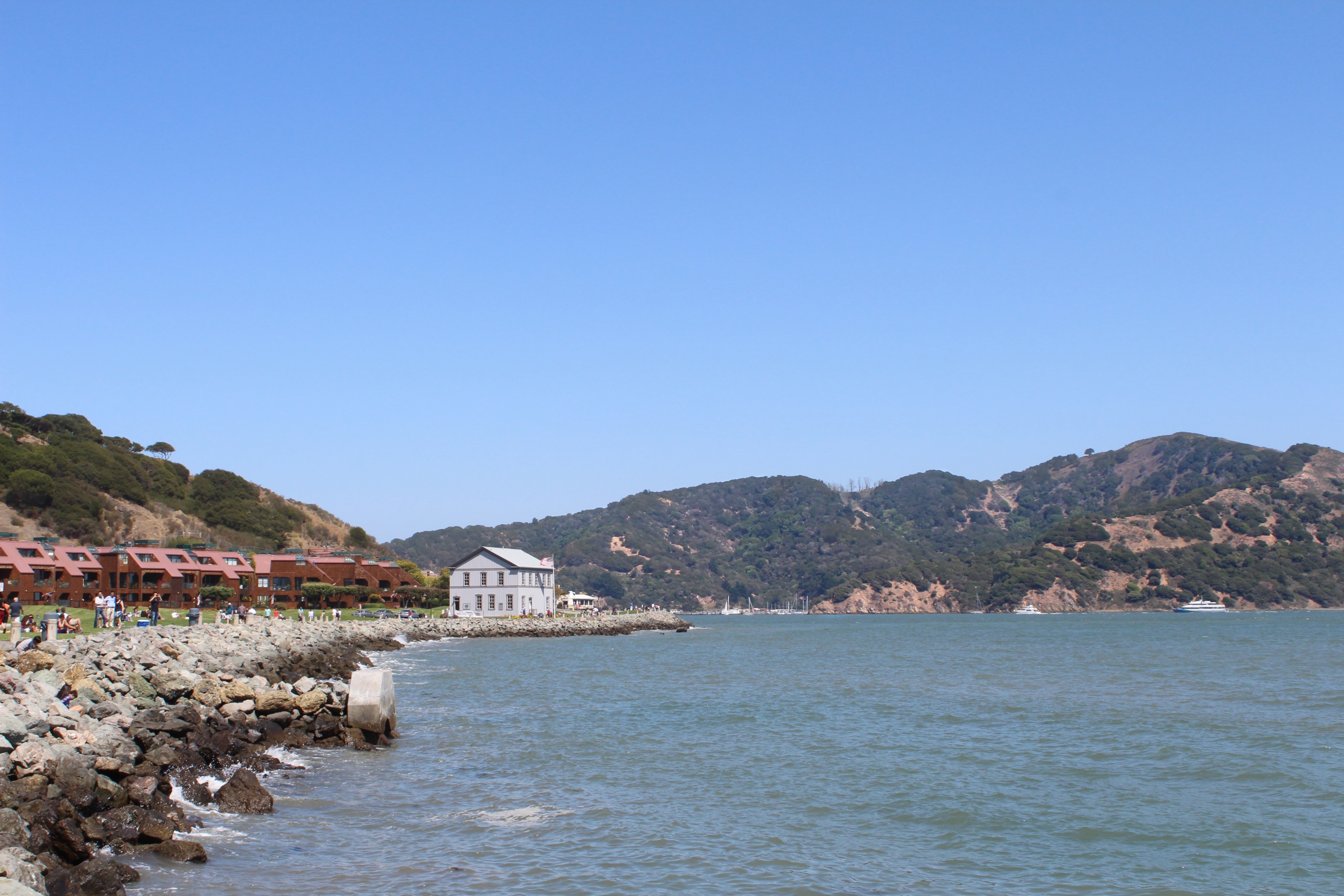 Rocky shoreline and calm bay water with red-roofed buildings and green hills.