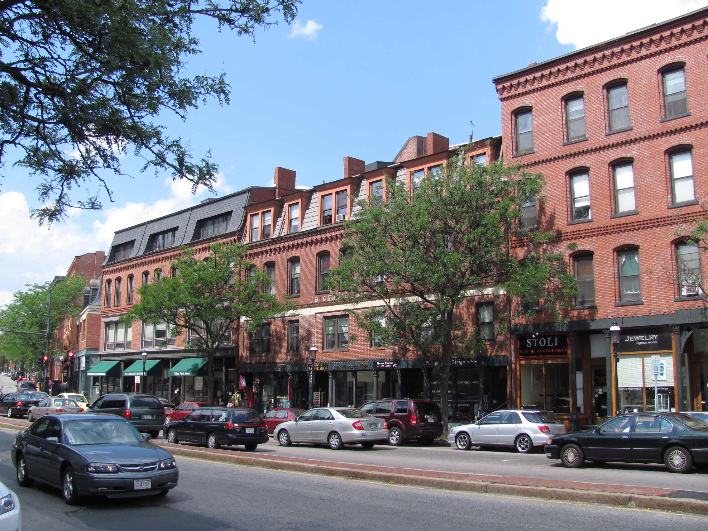 Row of multi-story red brick commercial buildings with ground-floor shops along a tree-lined street.