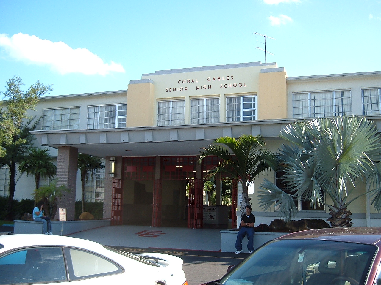 Art Deco-style school building entrance with palm trees and cars parked in front.
