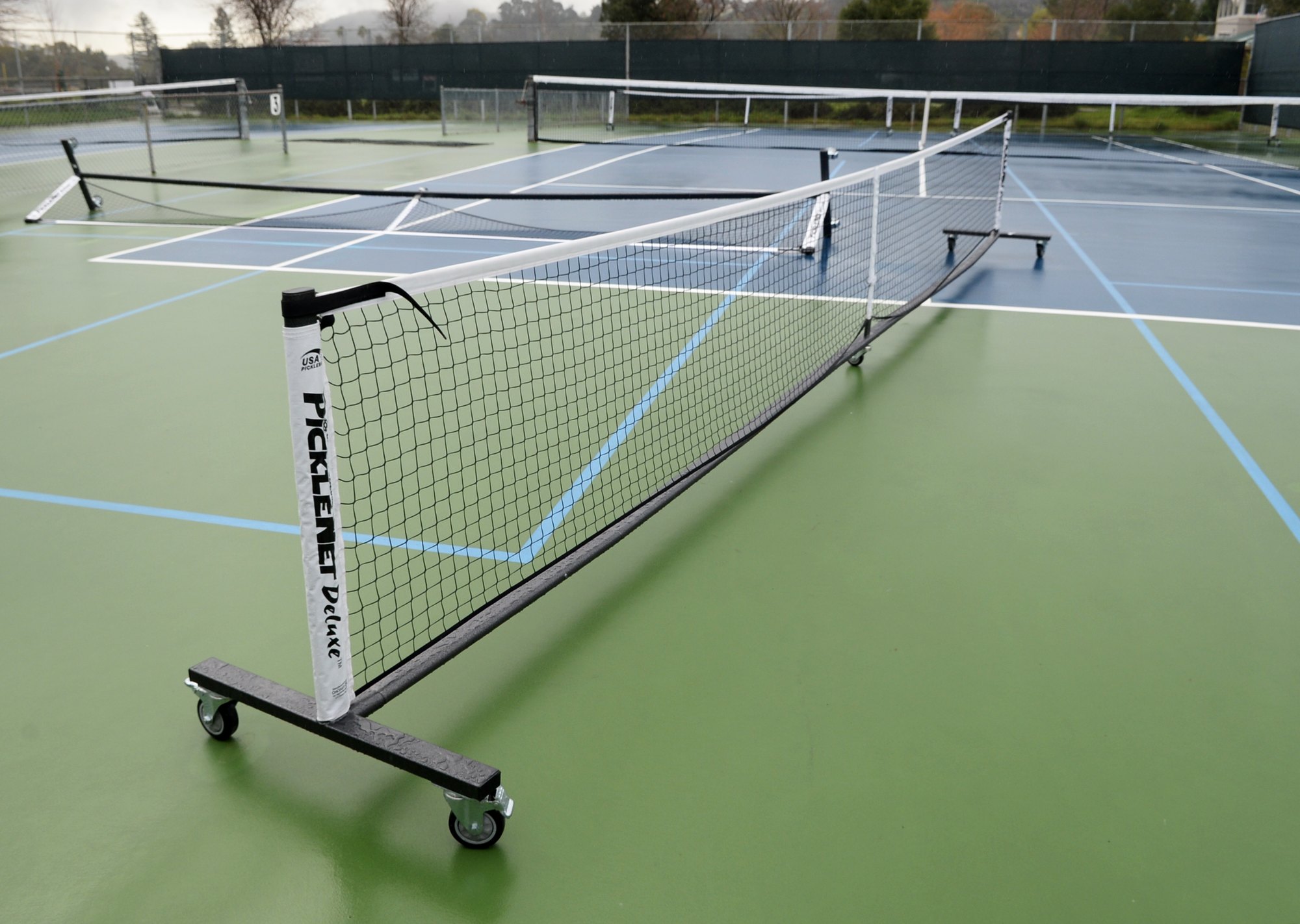 A portable pickleball net on wheels on a green multi-sport court with fencing in the background.