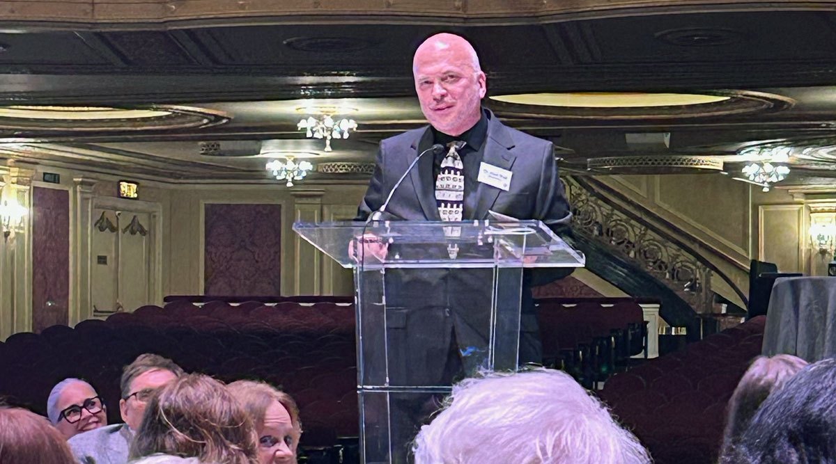 A man in a dark suit speaks at a clear acrylic podium in an ornate ballroom.