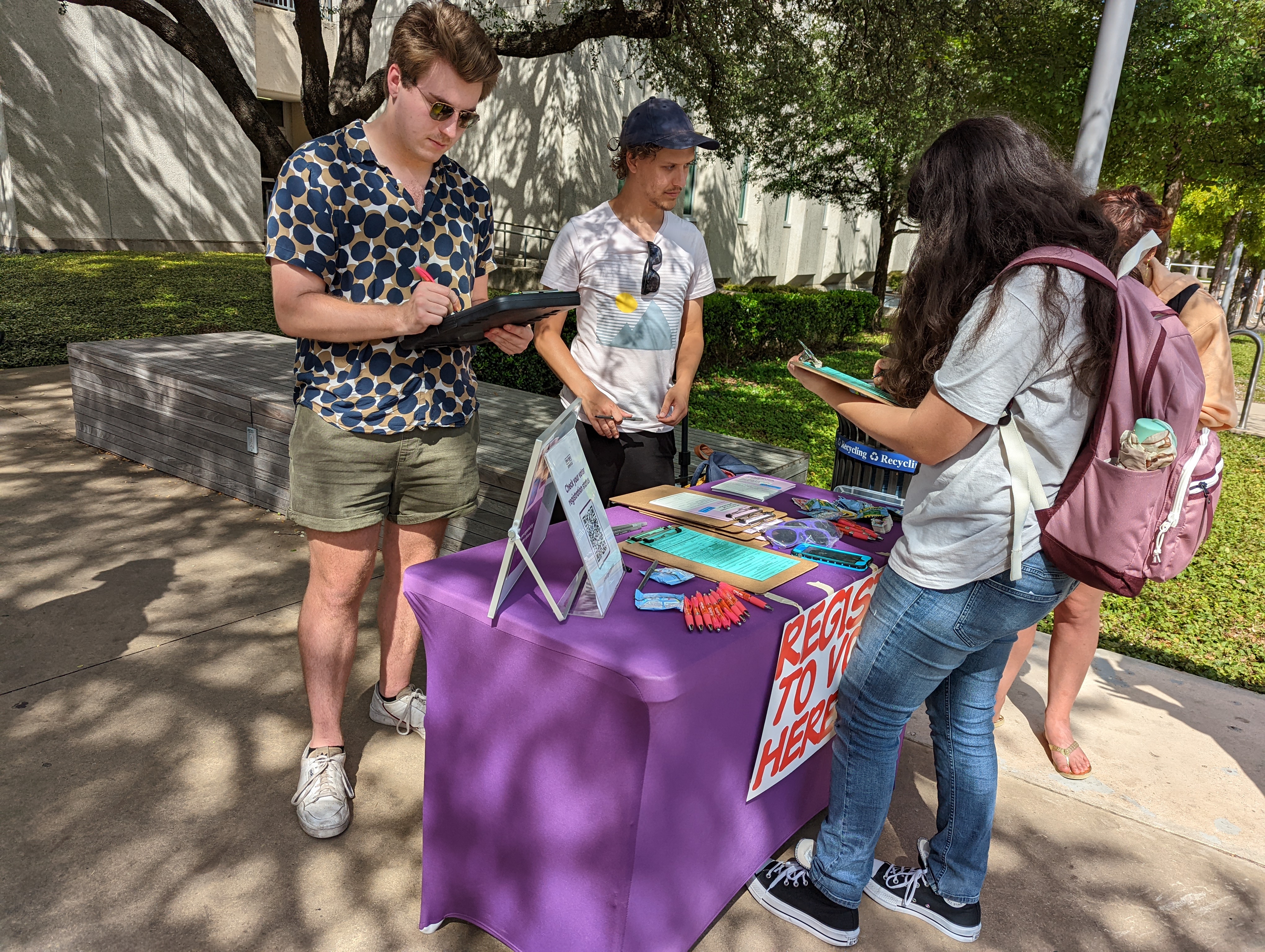 People at an outdoor voter registration table with a purple tablecloth and a sign reading Register to Vote Here.