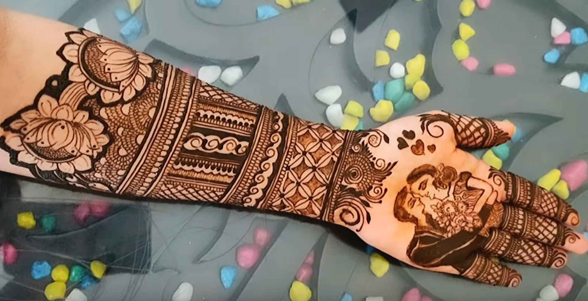 A hand and forearm adorned with elaborate henna designs featuring floral and geometric patterns with colorful confetti in the background.