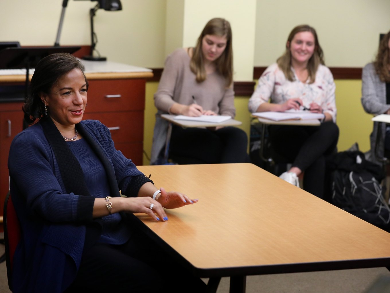 A woman in a dark blazer seated at a wooden table in a classroom, with students smiling behind her.