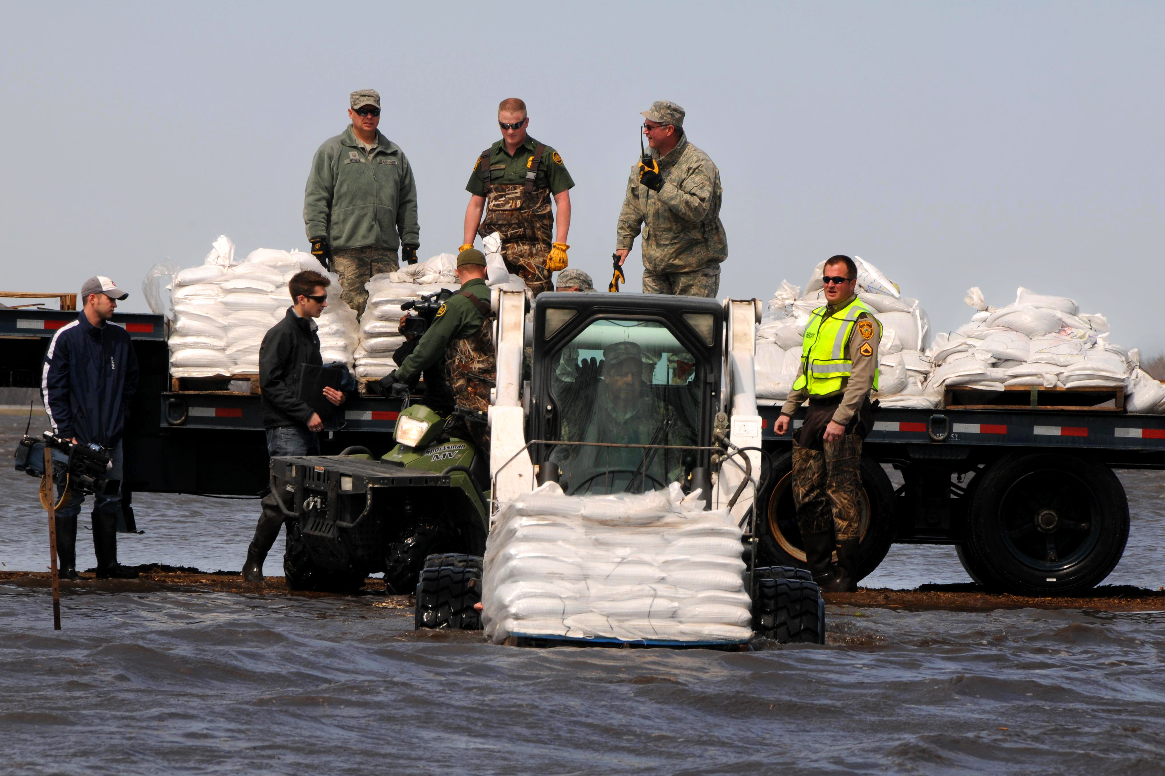 Workers load white sandbags from flatbed trucks using a skid-steer loader.