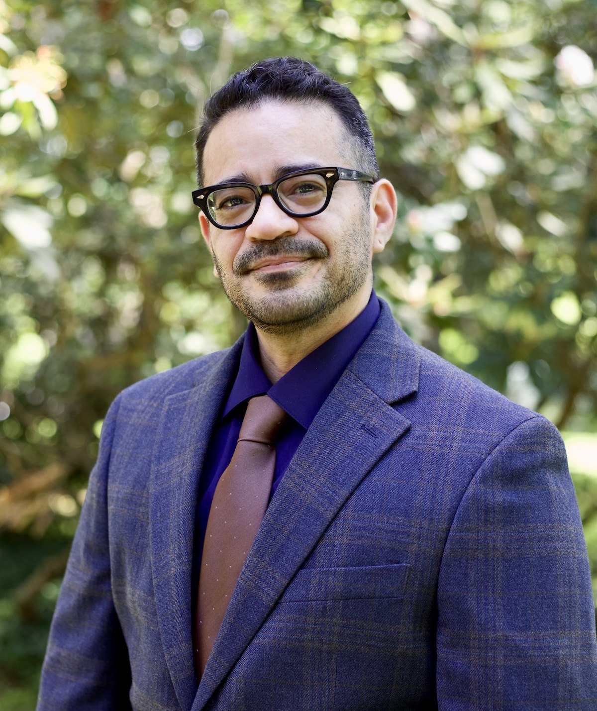 Man in blue plaid blazer and glasses poses outdoors in front of green foliage.
