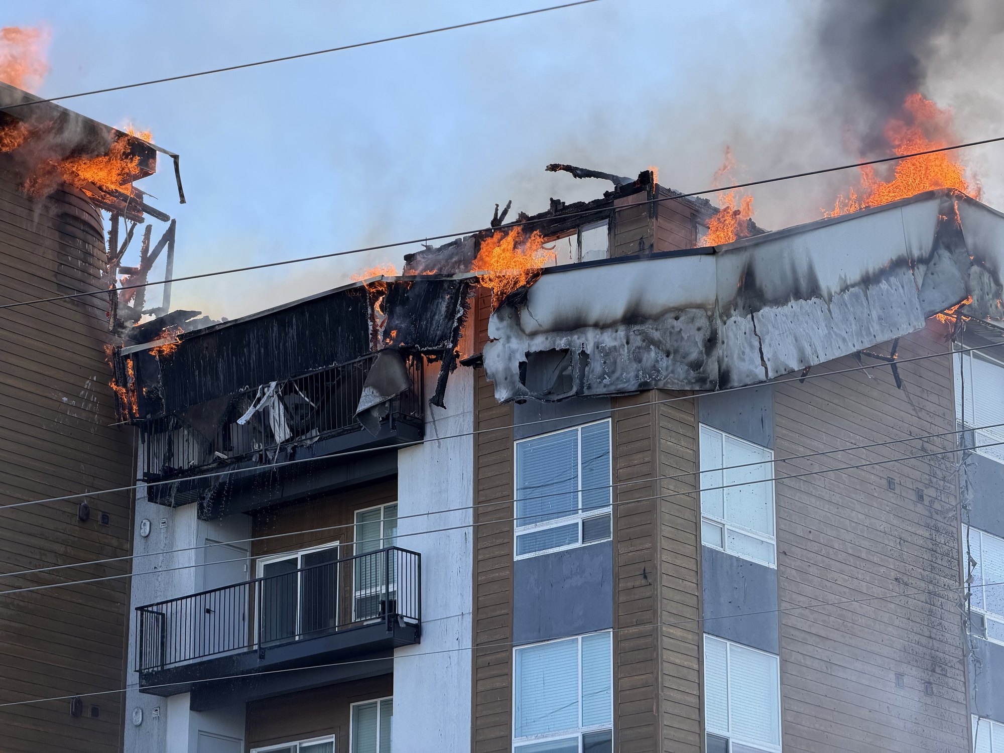 Flames and charred roofing materials burning across the top of a multi-story apartment building.