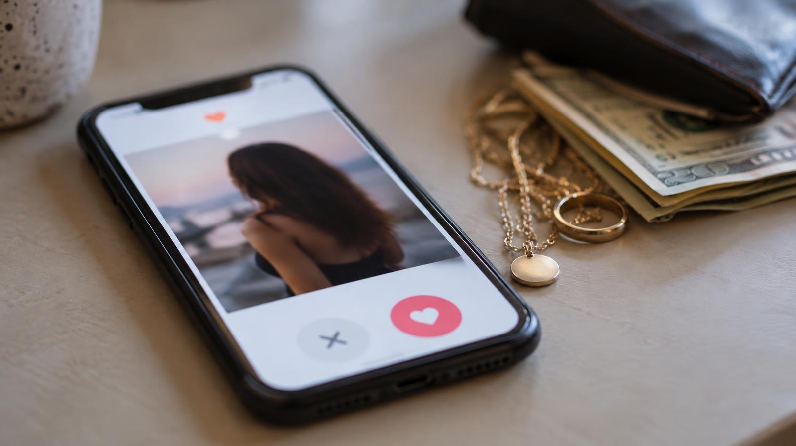 Smartphone with dating app screen next to jewelry and folded cash on a surface.