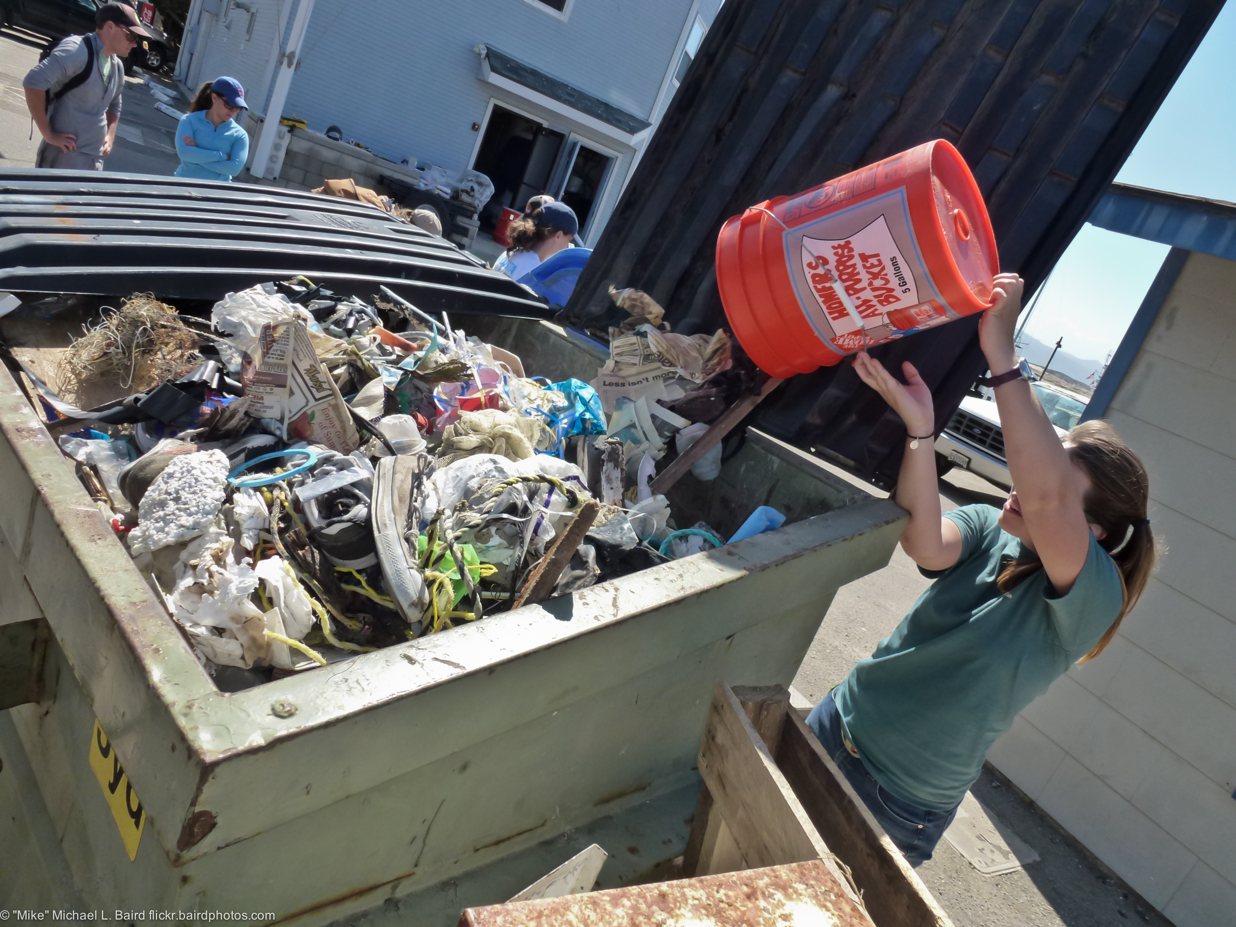 Volunteer hoisting an orange bucket to dump trash into an overflowing bin at a beach cleanup event.
