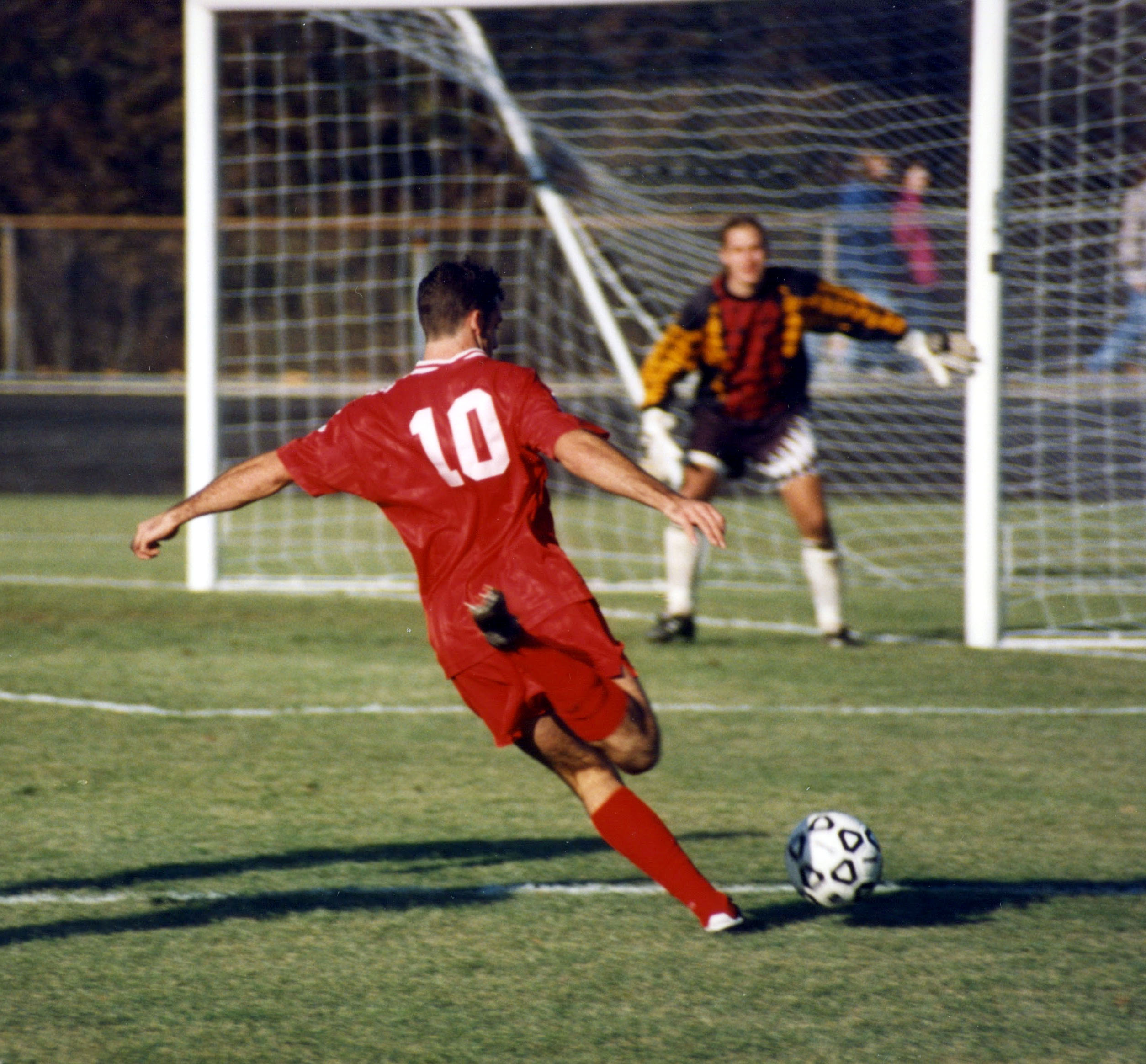 A soccer player in a red jersey drives toward the goal as a goalkeeper defends.