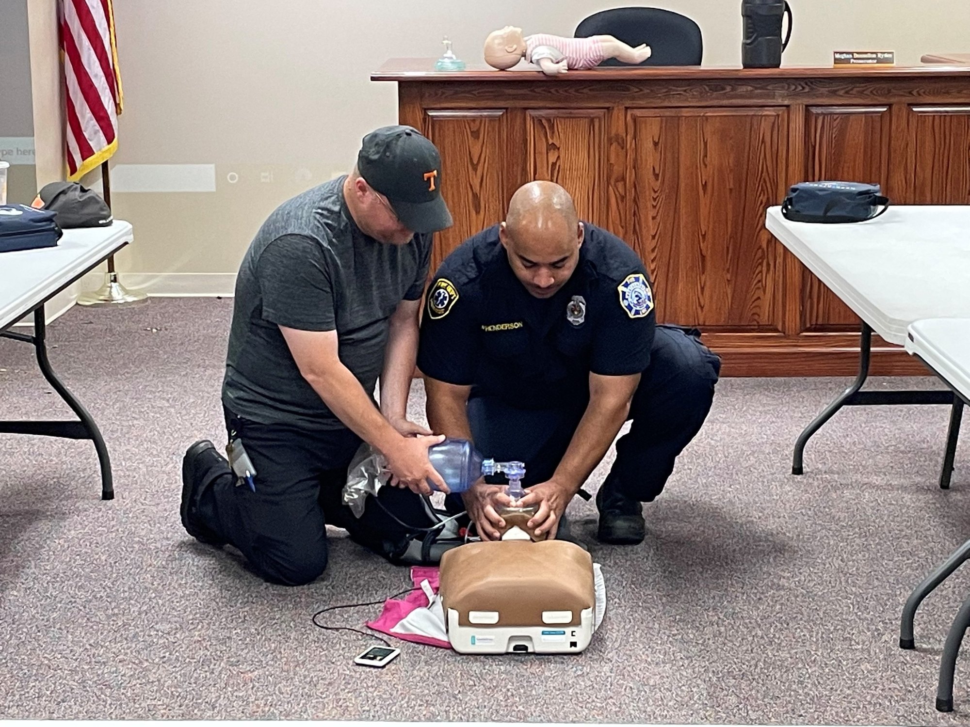 Two men kneel on a floor practicing CPR on a training manikin with an AED device nearby.