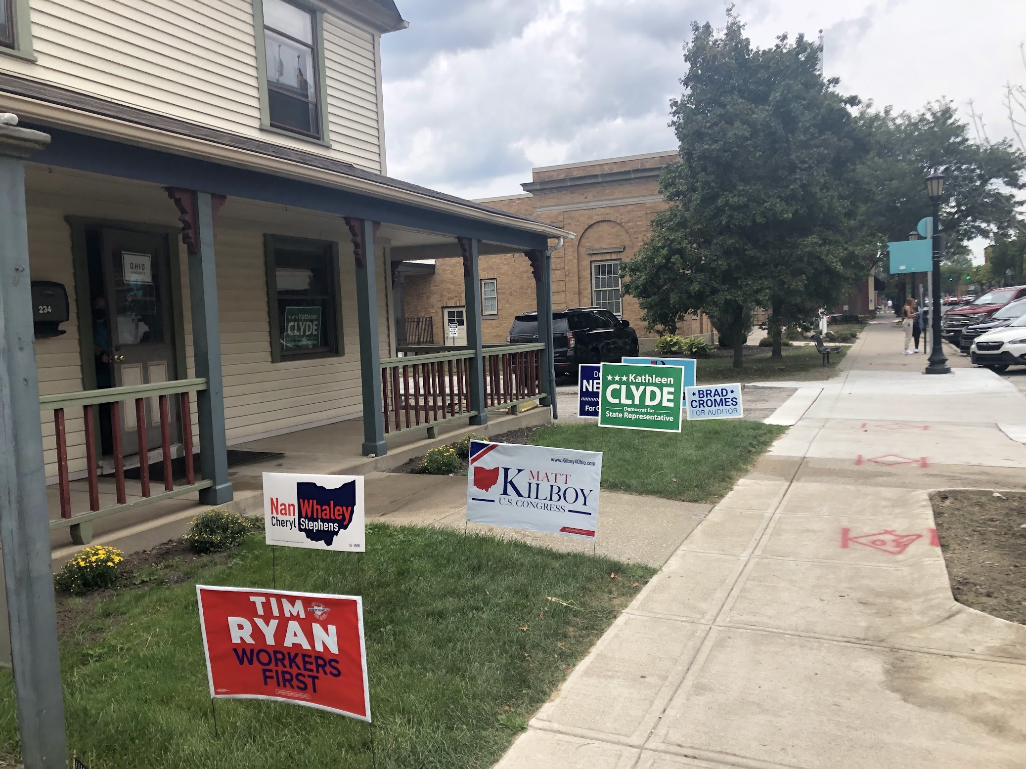 Multiple colorful political campaign yard signs lined along a sidewalk.