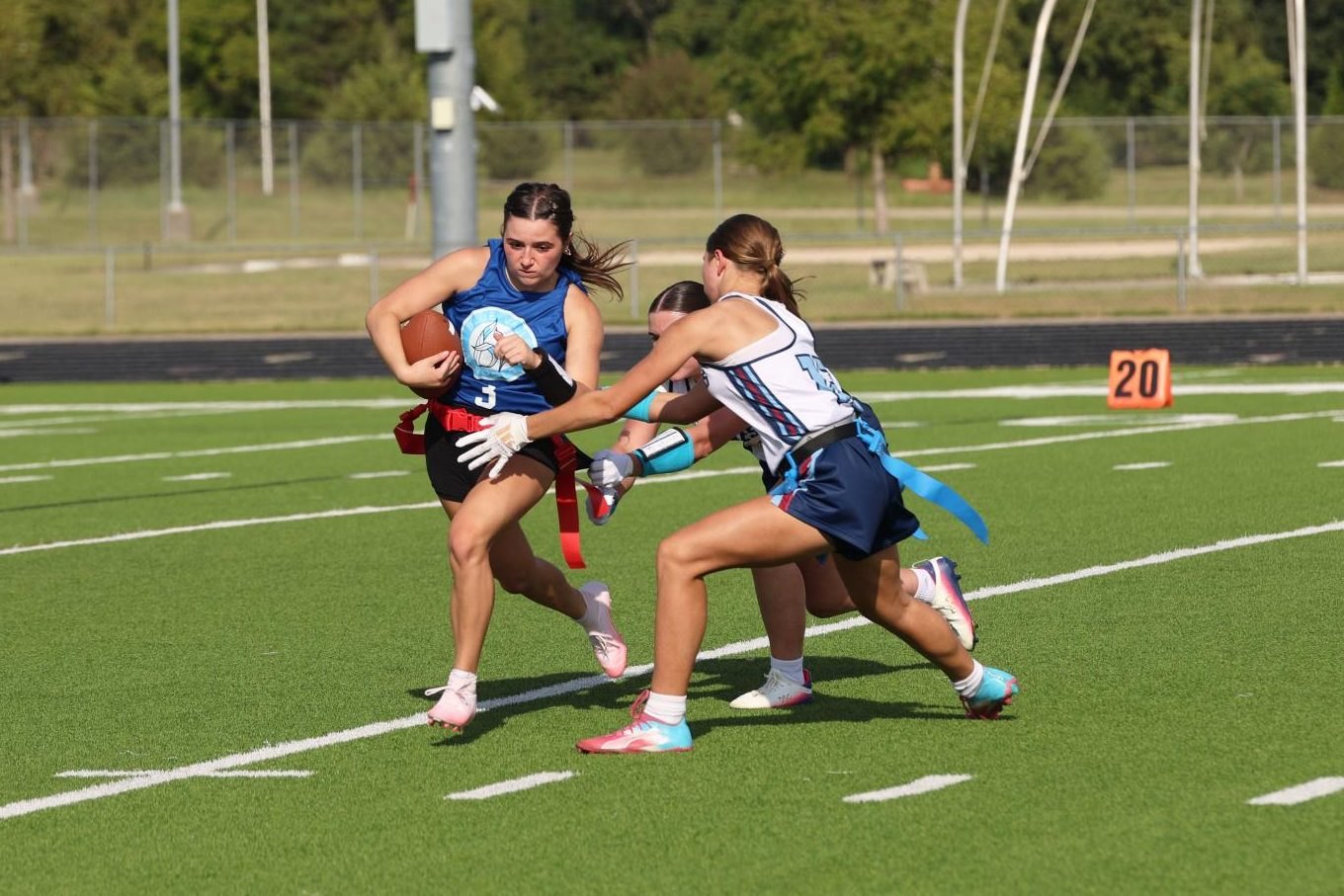 Female flag football player in blue jersey runs with the ball as a defender reaches for her flag.