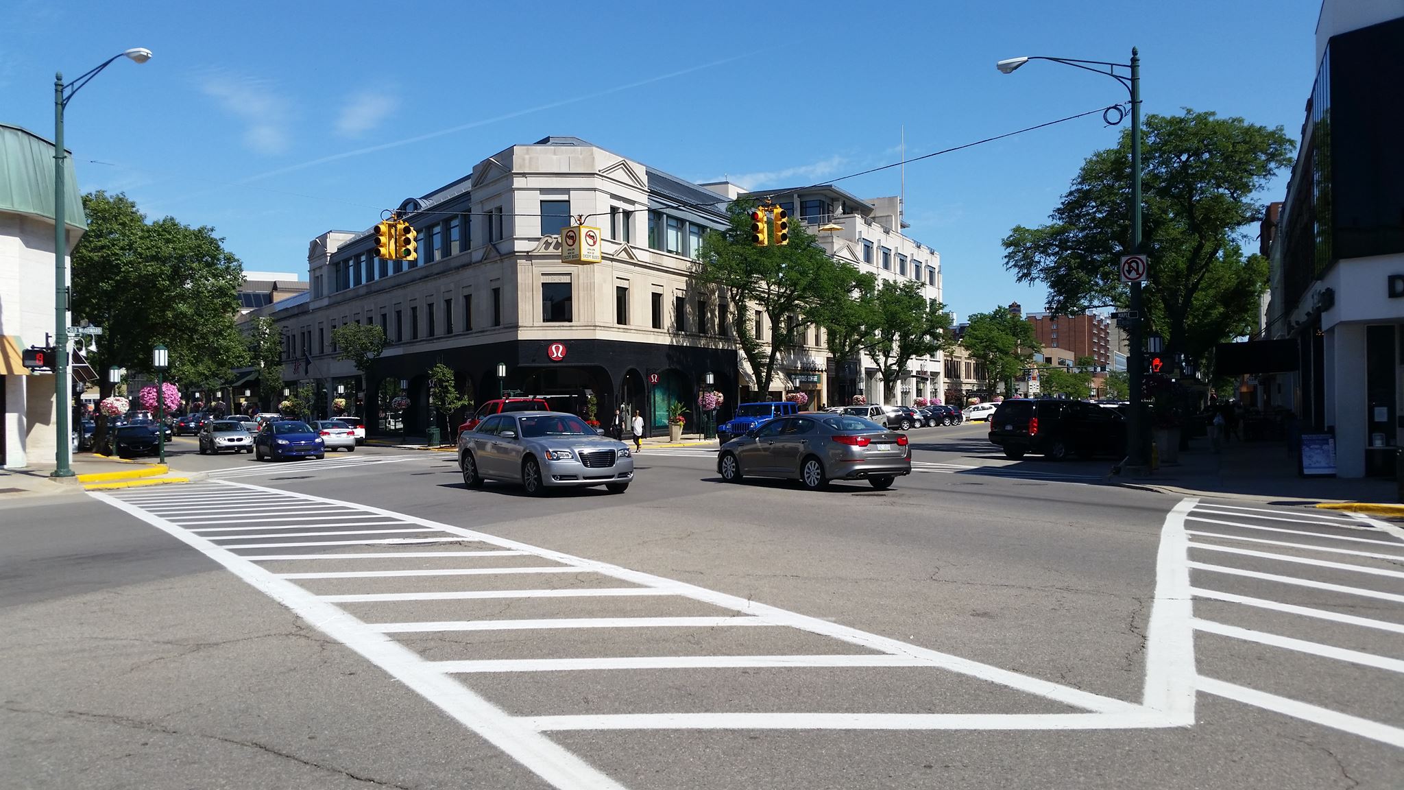 Busy downtown intersection with vehicles, traffic signals, and brick commercial buildings on a sunny day.