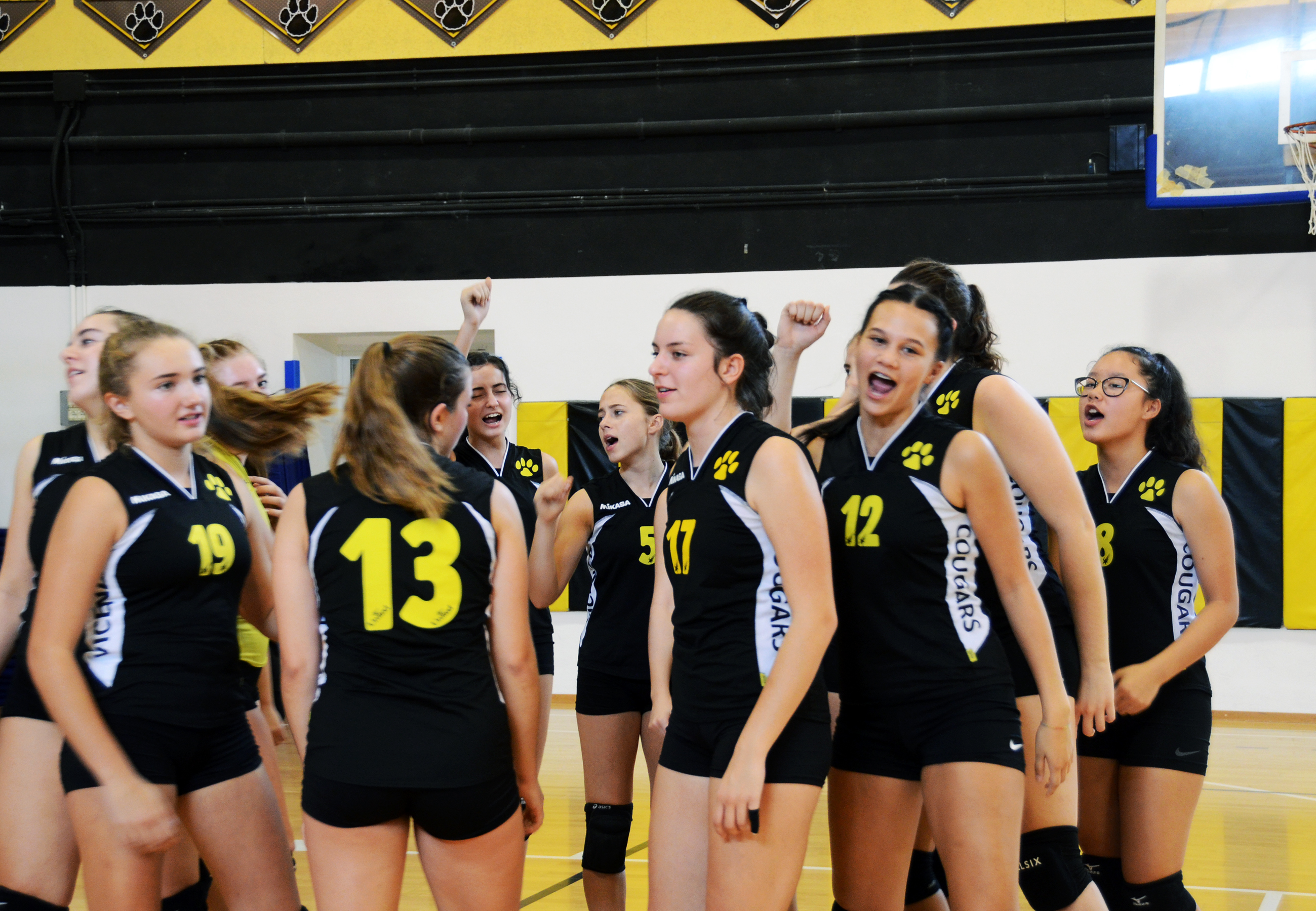 Female volleyball players in dark uniforms celebrate together in a gymnasium.