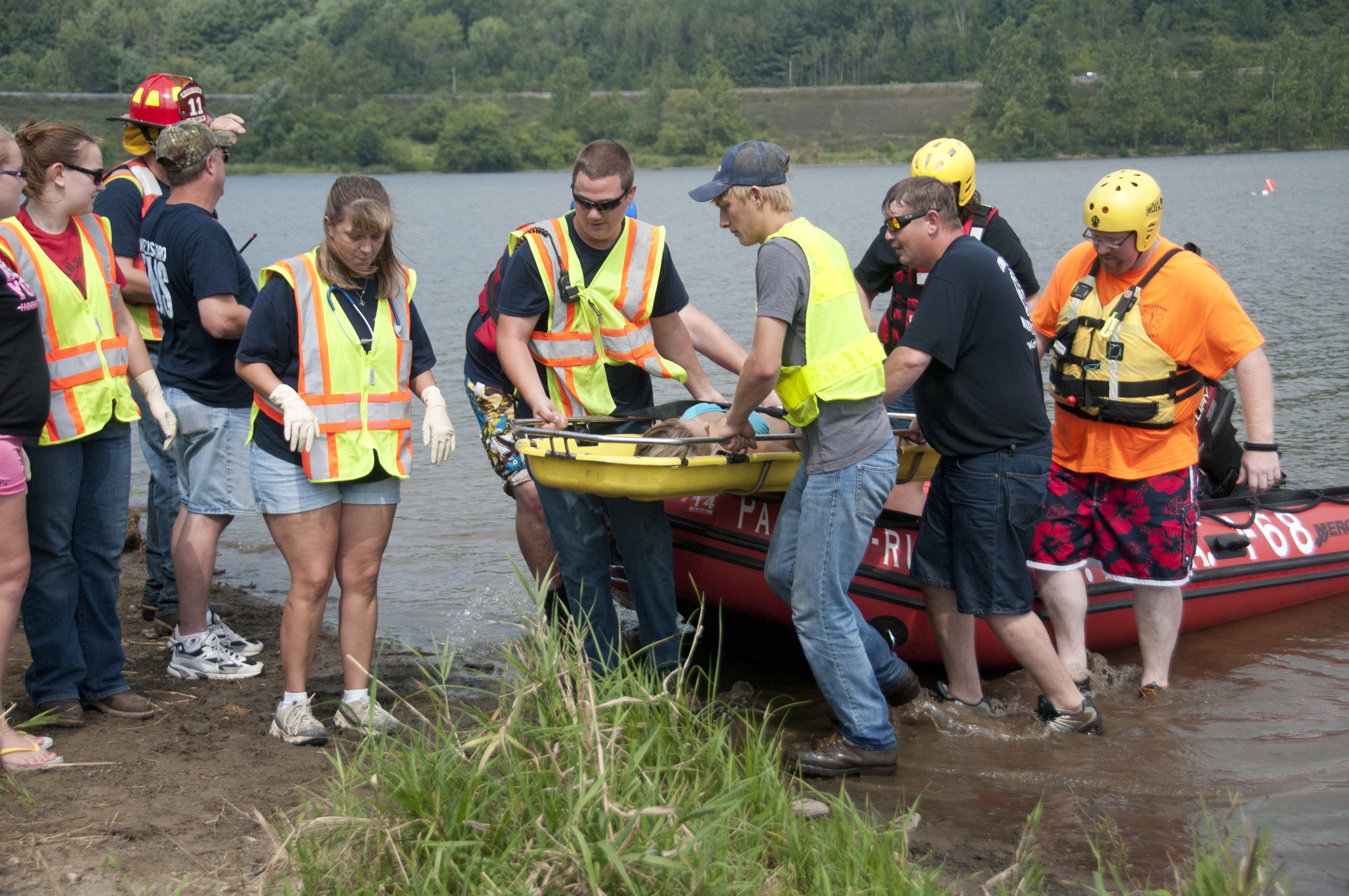 Emergency responders in safety vests carry a yellow stretcher from a red inflatable rescue boat at a lake's edge.