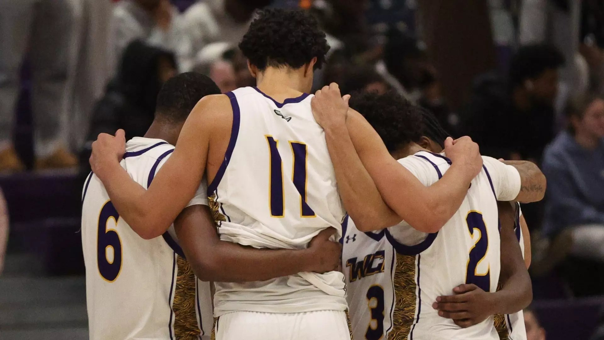 Five basketball players in white and purple uniforms huddle with arms around each other on an indoor court.