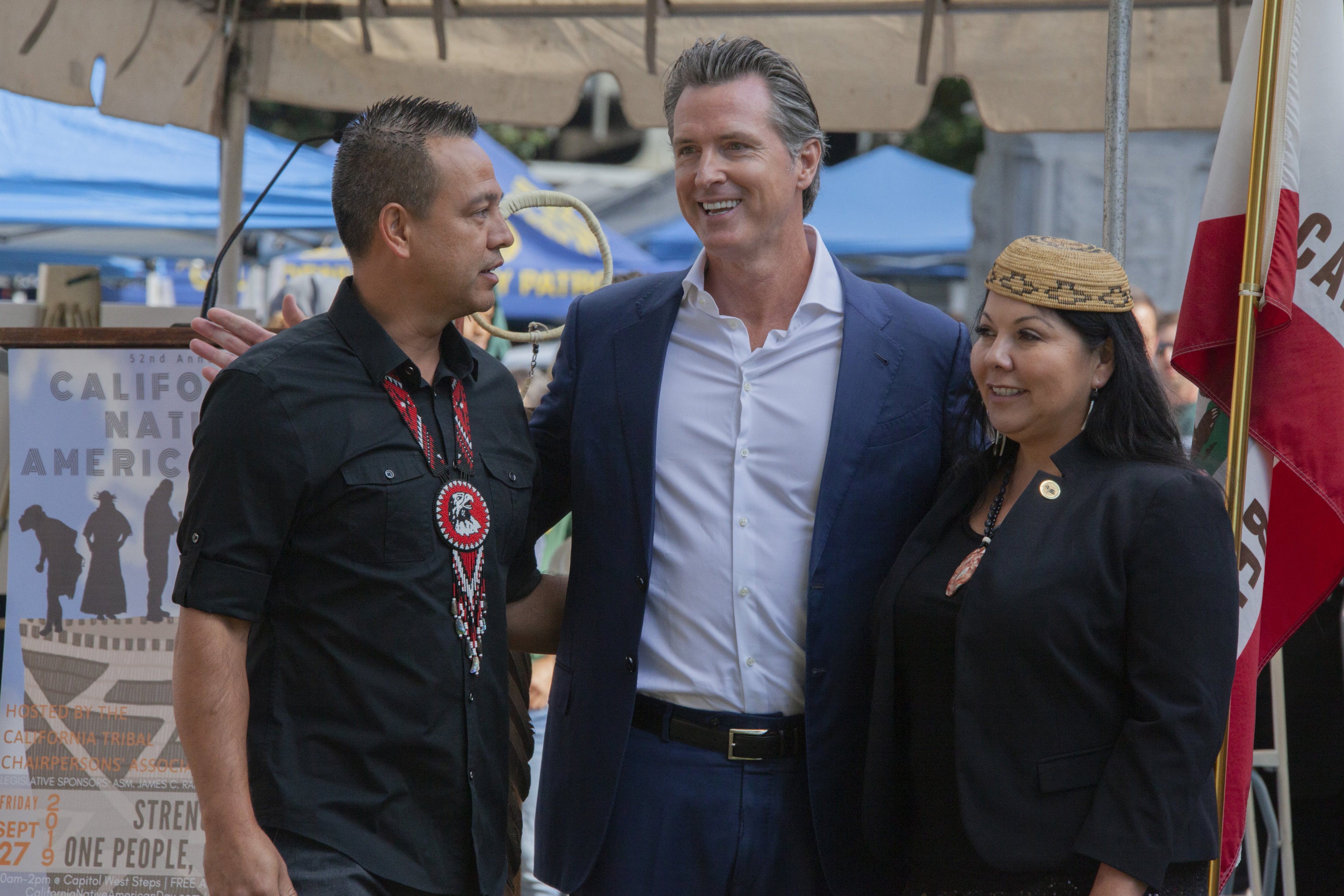 Three people pose outdoors at a ceremony with a California state flag and Native American Day banner visible.