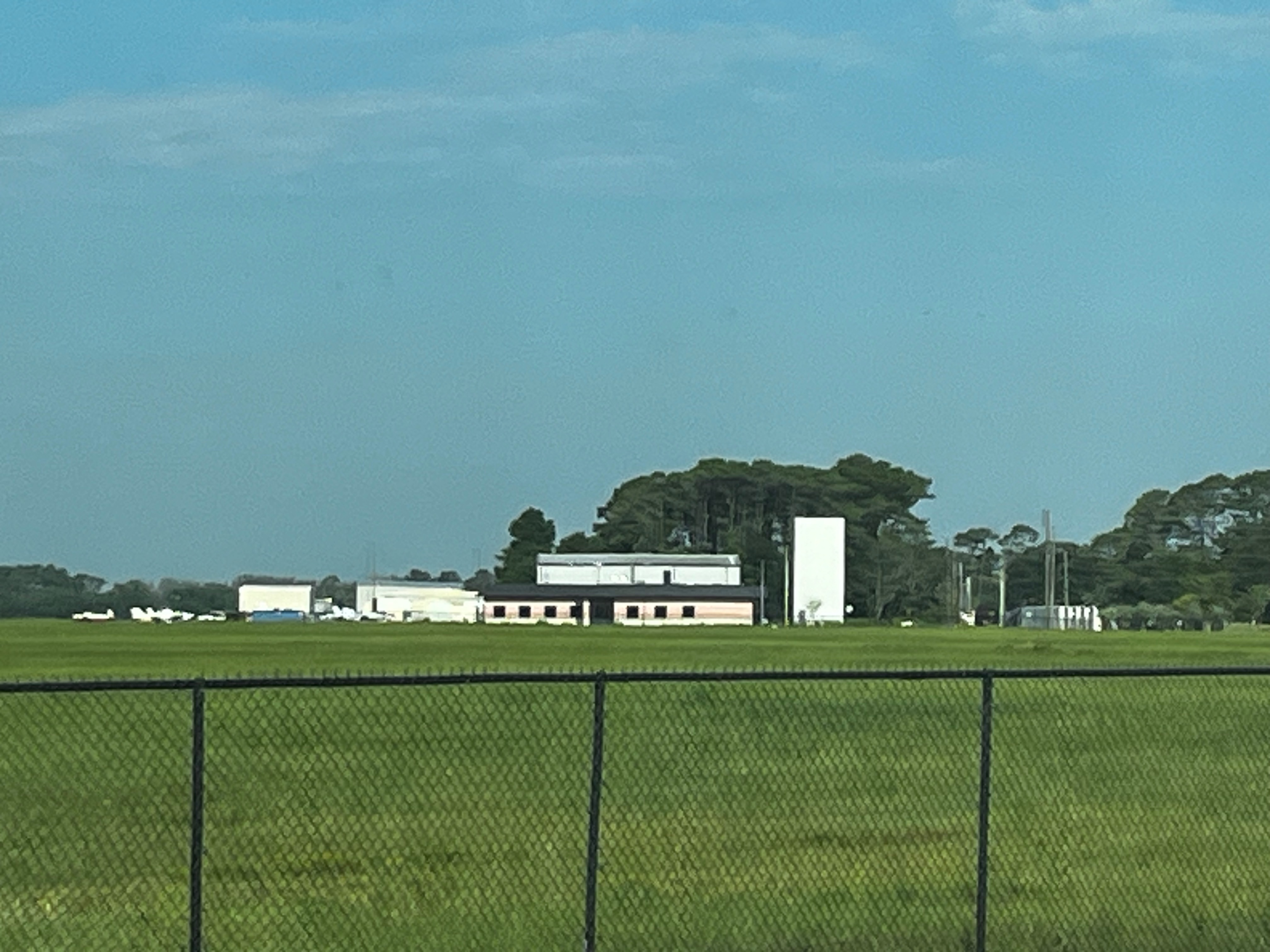 Airport hangars and buildings viewed across a flat grassy airfield under a blue sky.