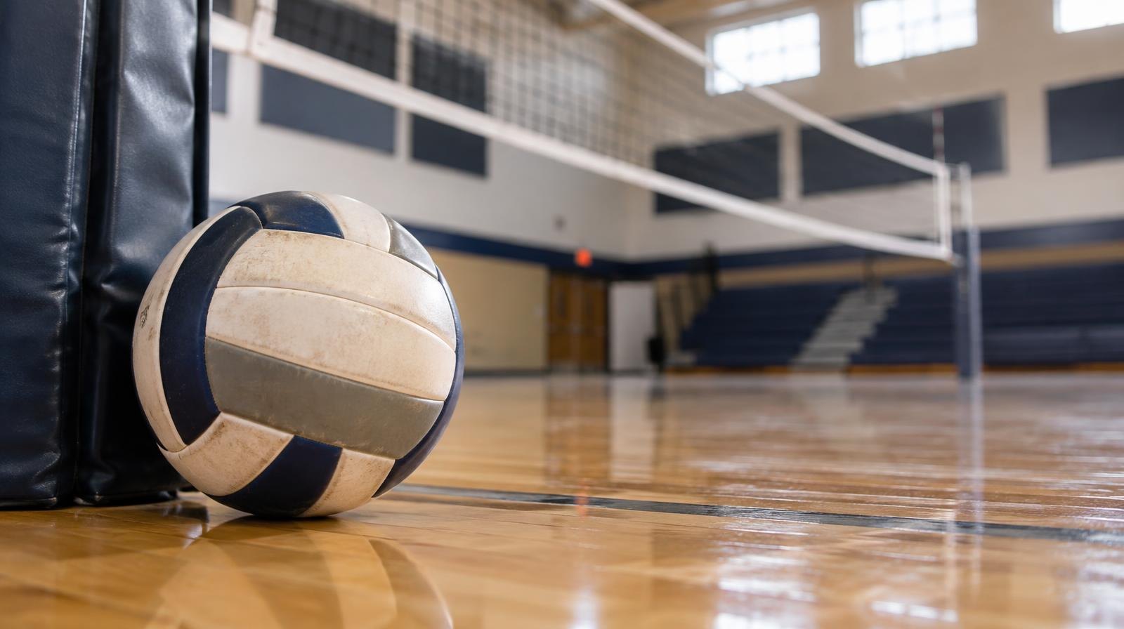 A volleyball resting on a hardwood gym floor beside a net post.
