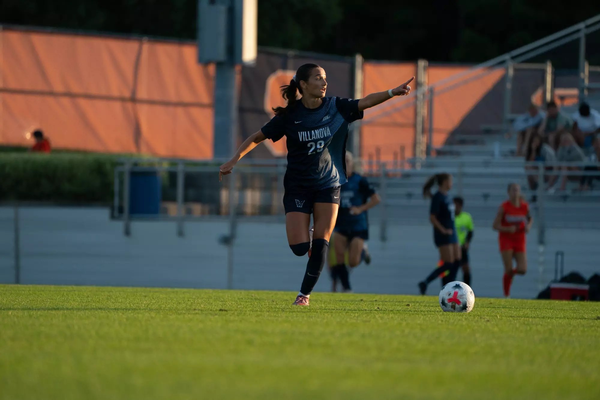 A female soccer player in a dark Villanova number 29 jersey runs on a grass field with a ball nearby.