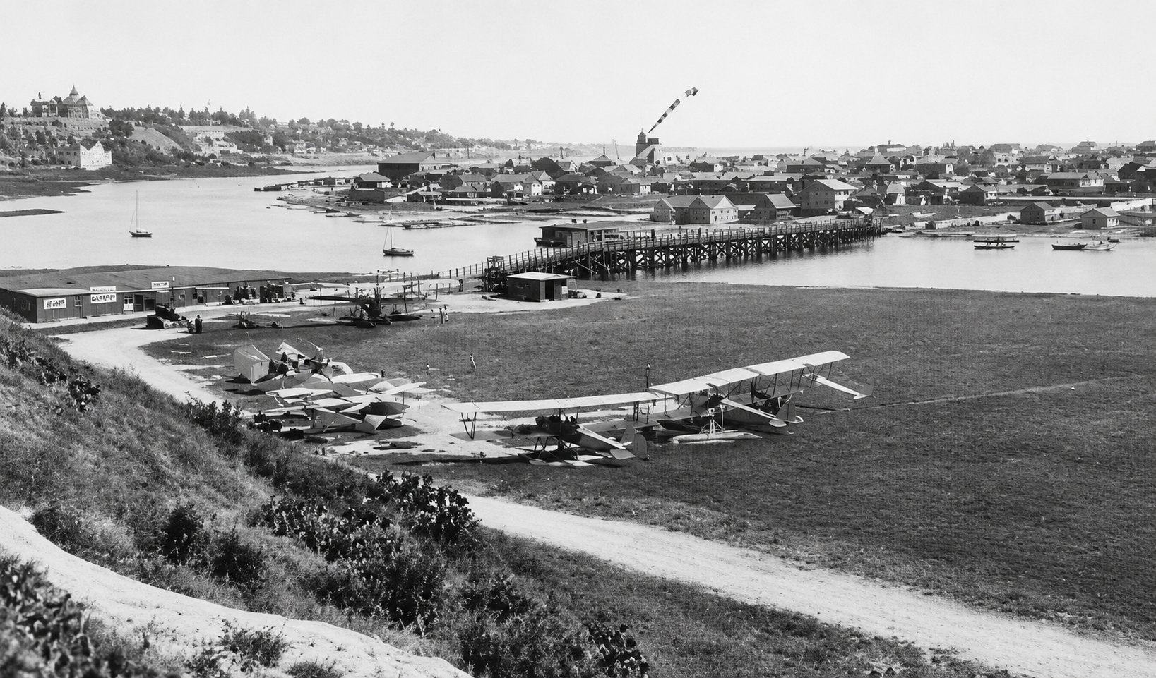Historic black-and-white photo of biplanes on a grassy coastal airstrip with a pier and harbor in the background.