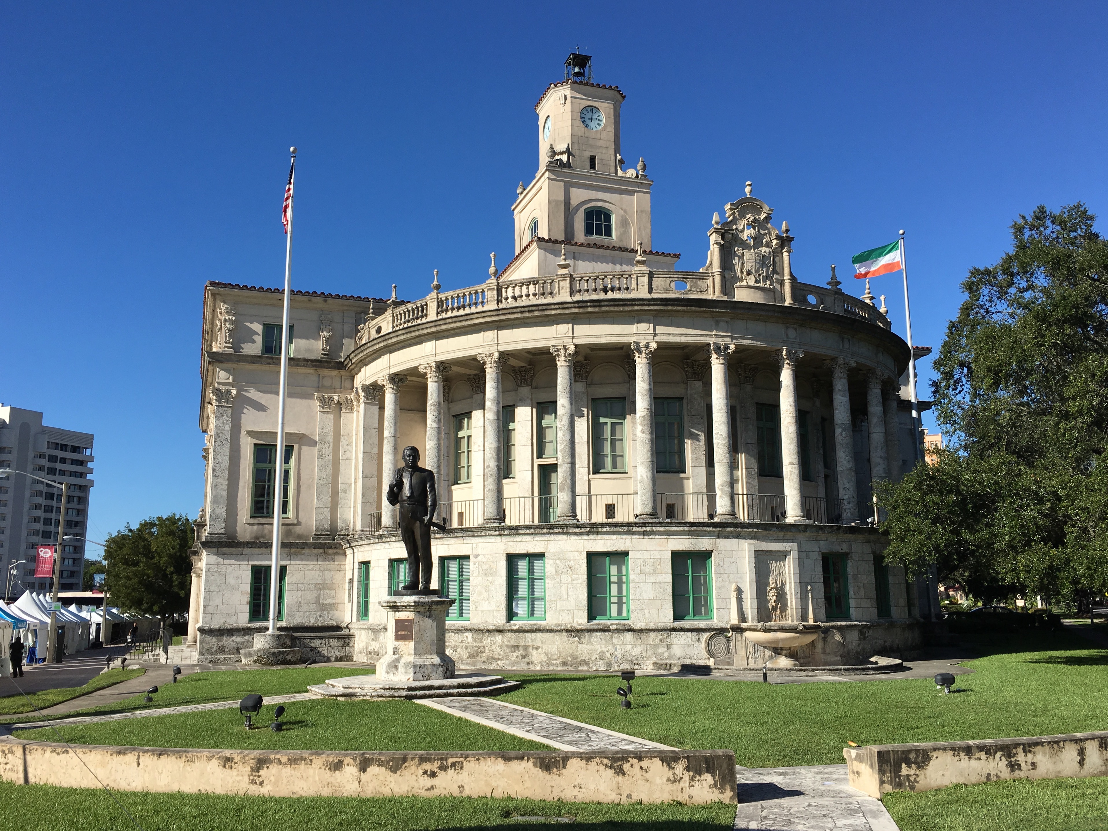Neoclassical government building with clock tower, colonnade, bronze statue, and two flagpoles under blue sky.