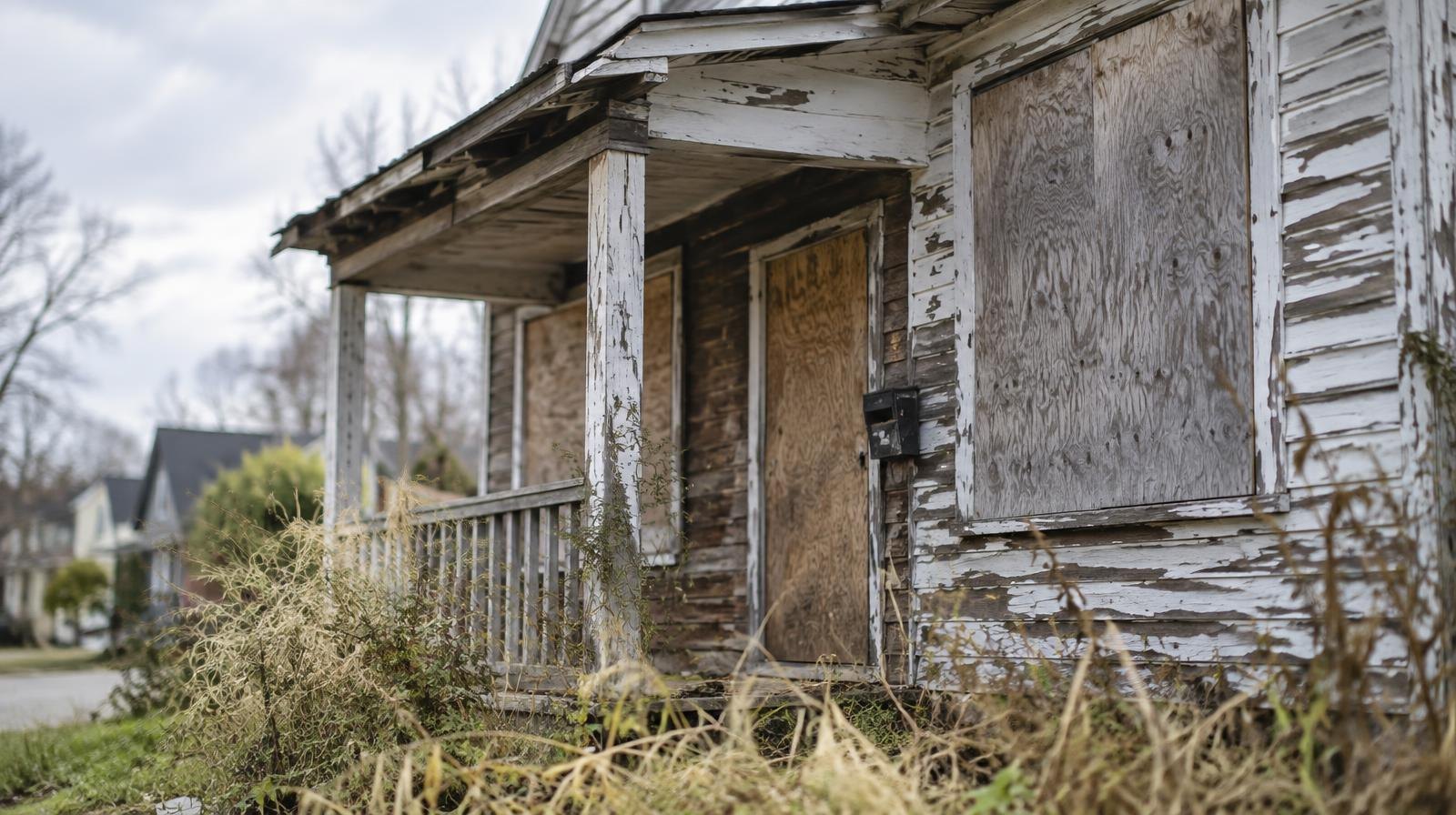 Weathered boarded-up abandoned house with peeling paint and overgrown weeds.