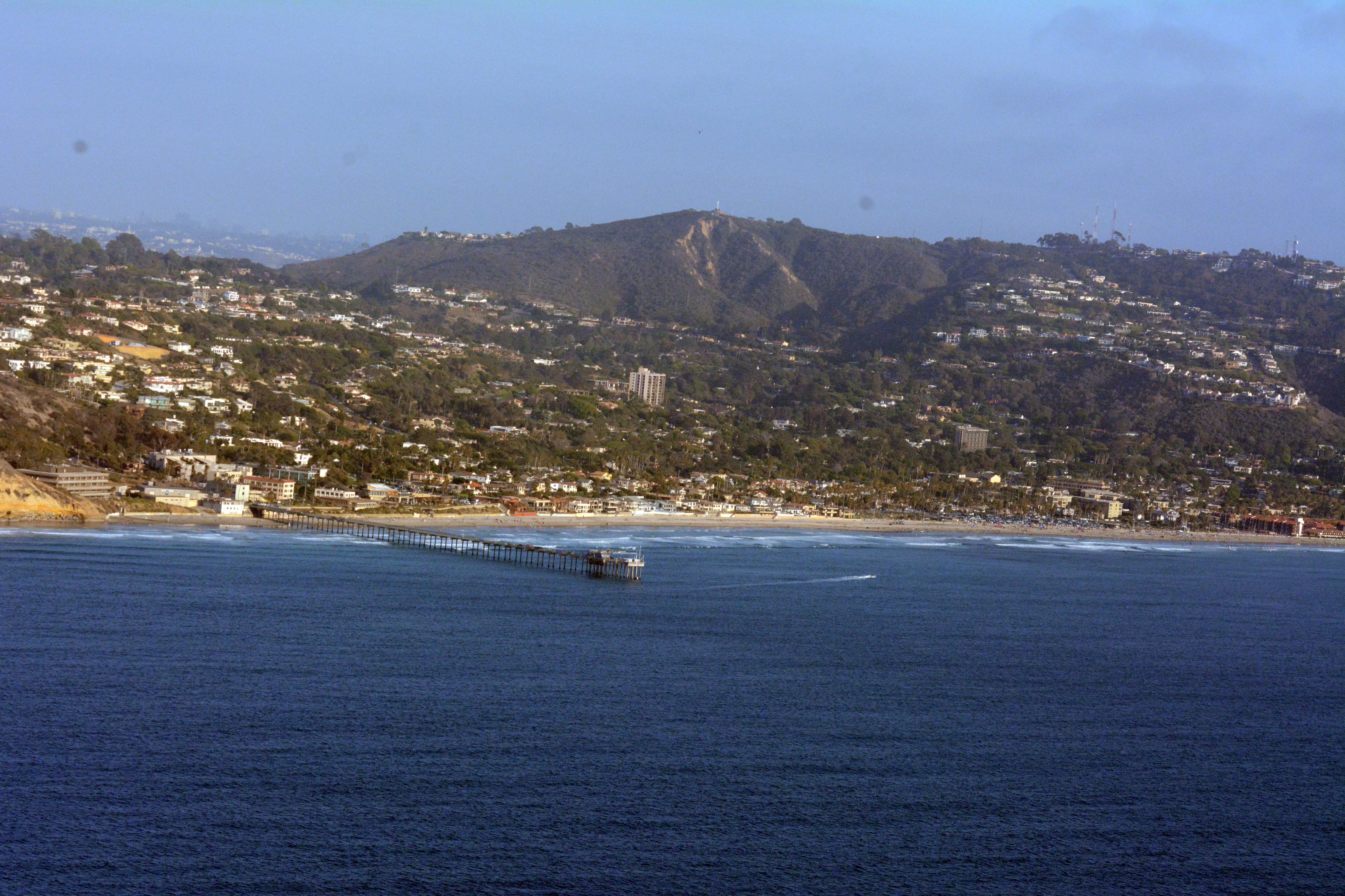 Aerial view of a long research pier extending into the blue ocean with a sandy beach and hillside community behind.